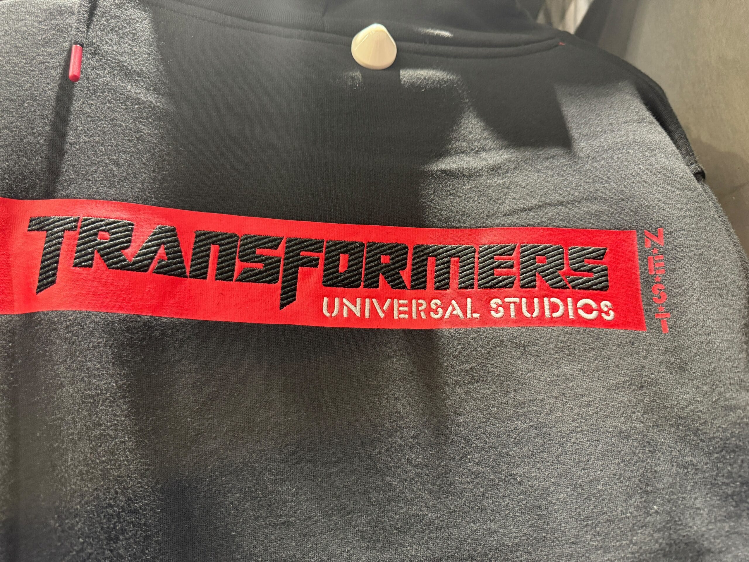 Black hoodie with a red stripe reading "TRANSFORMERS UNIVERSAL STUDIOS NEST" in bold, stylized text.