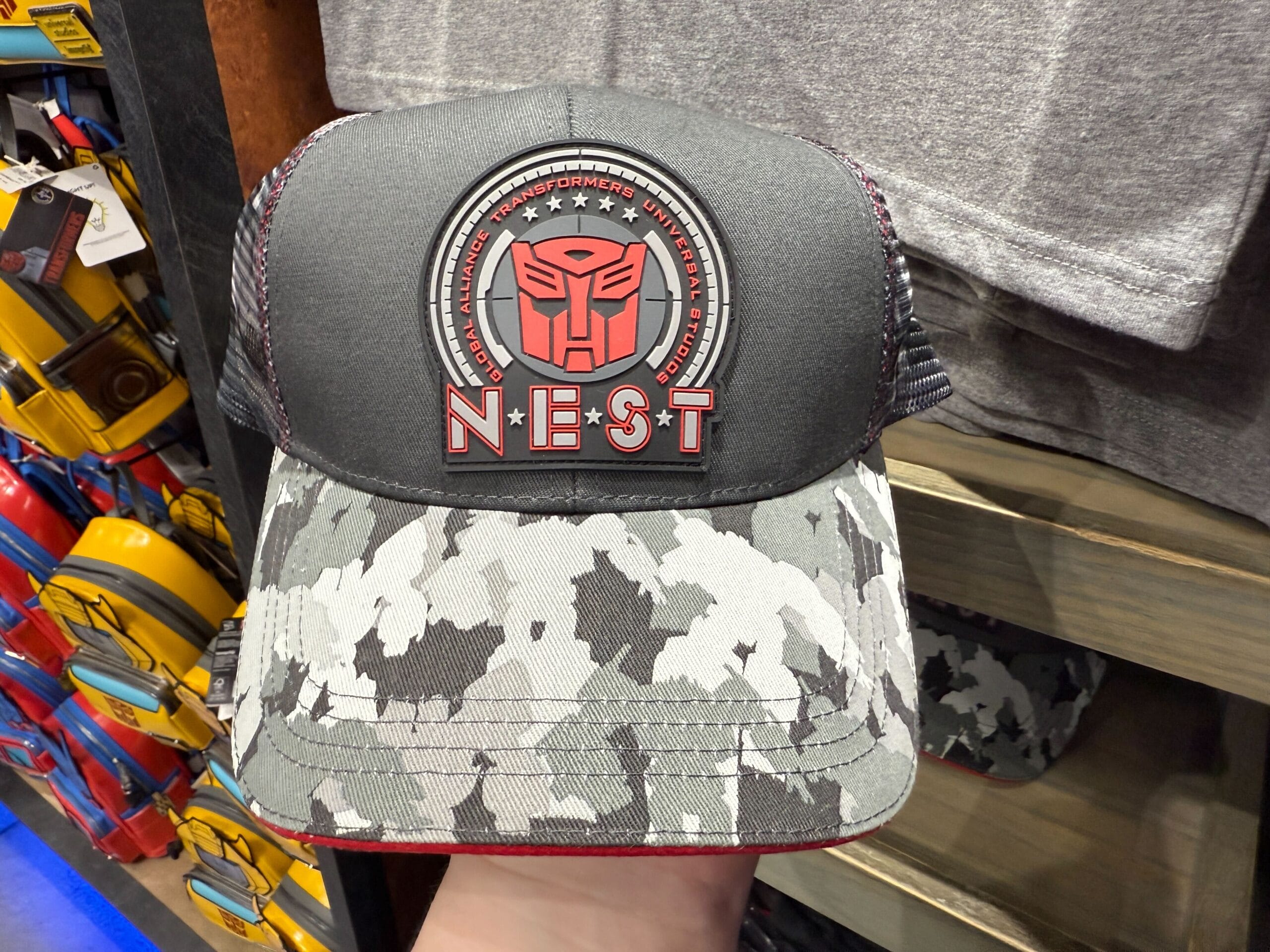 A black and gray camo cap featuring a red Transformers NEST logo and emblem on the front panel.
