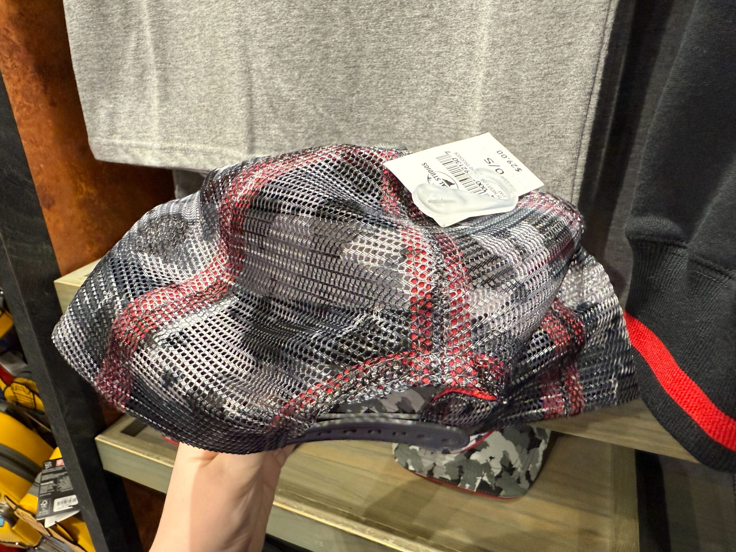 Hand holding a mesh baseball cap with a camouflage and red plaid pattern, displayed in a clothing store.