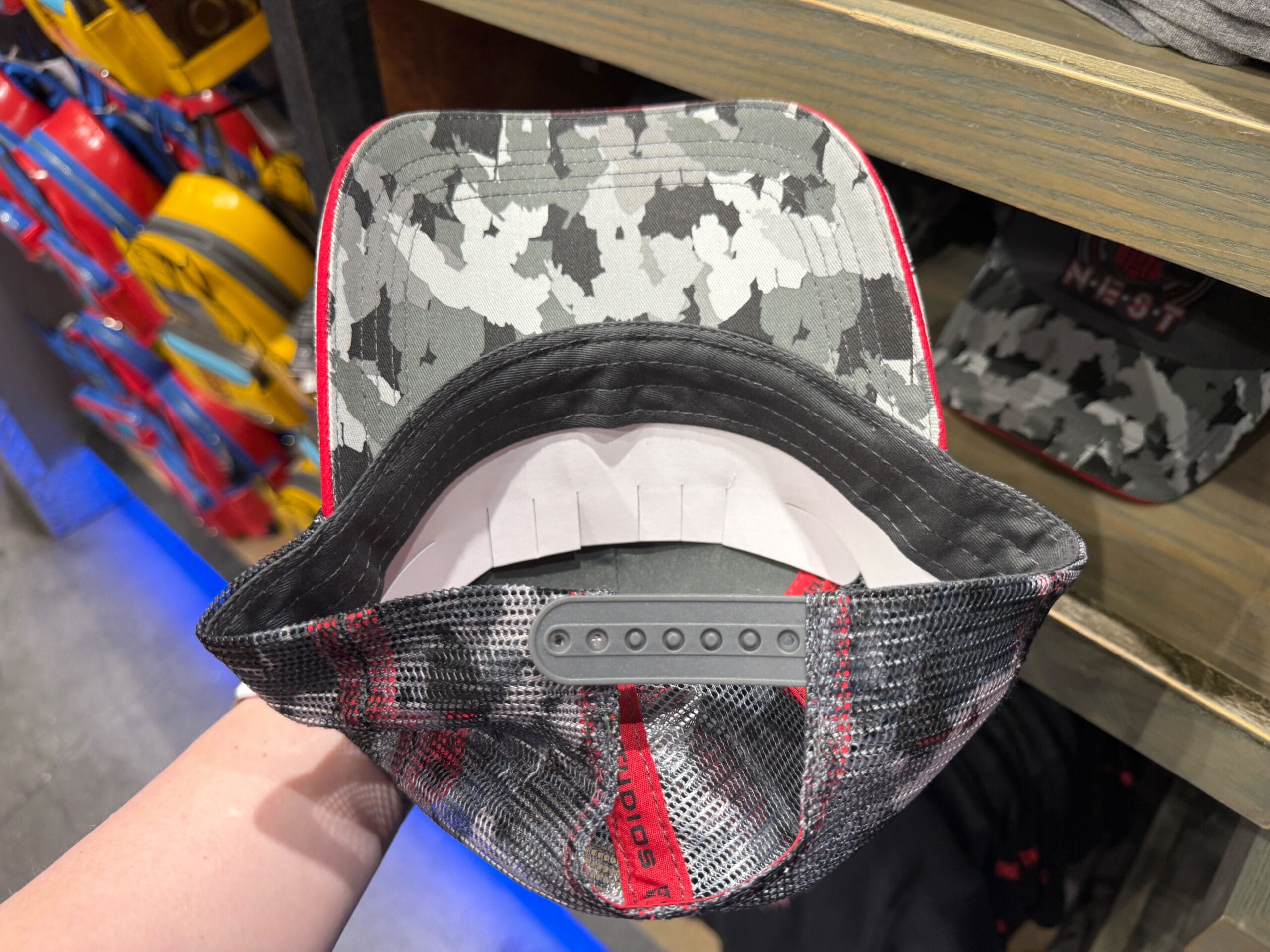 A hand holds a gray camo snapback hat with a red edge, showing the hat’s underside and adjustable strap.
