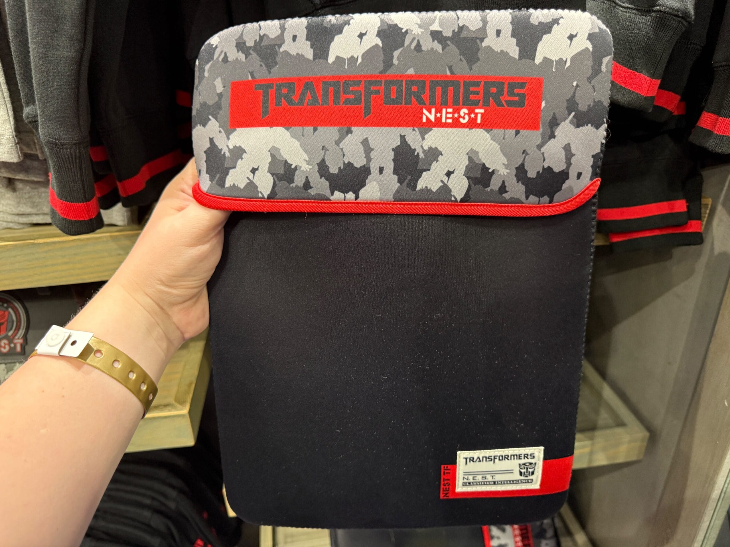 A hand holds a "Transformers N.E.S.T." themed tablet sleeve with a gray camo pattern and red accents.