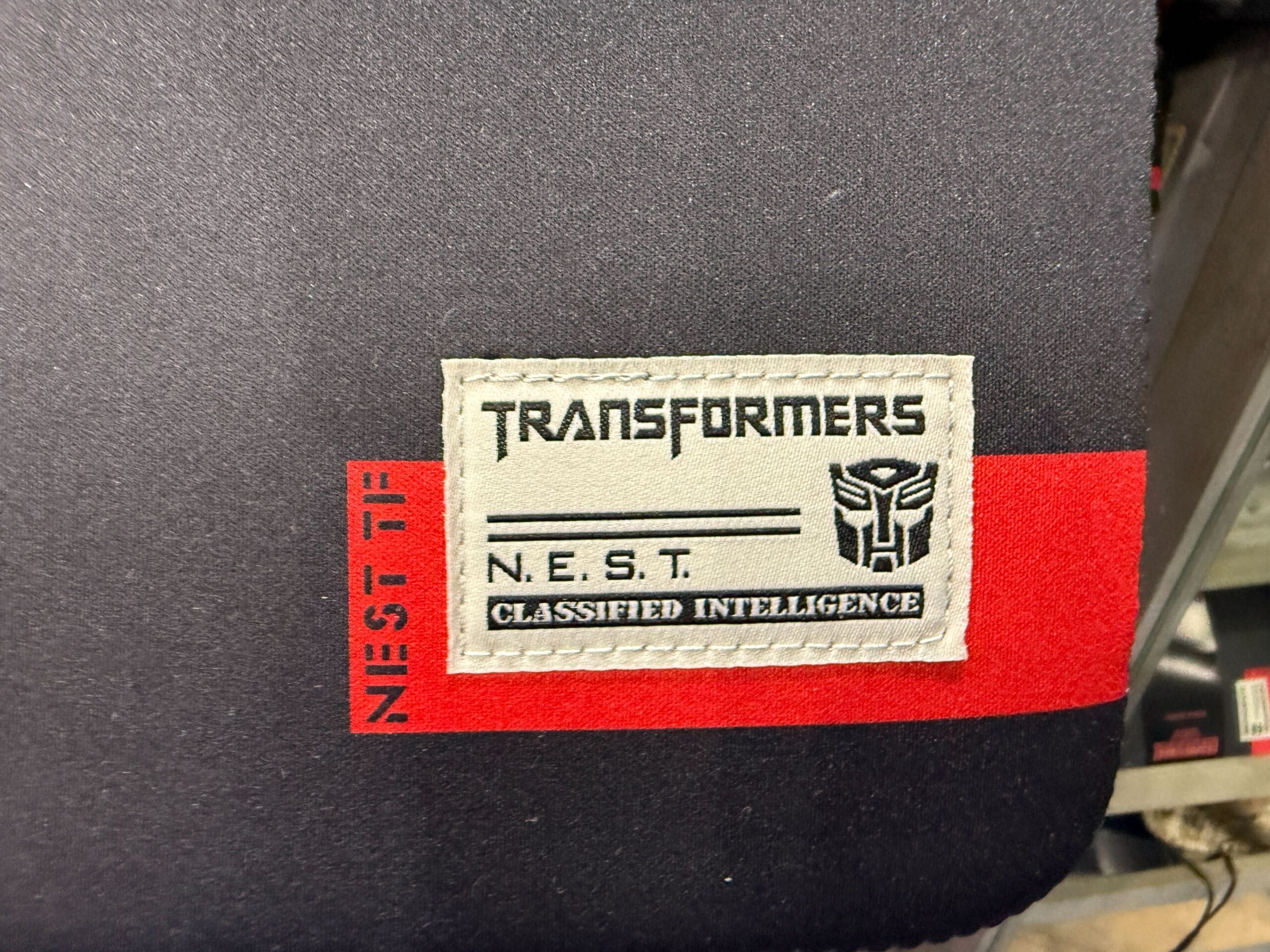 A patch reads "TRANSFORMERS N.E.S.T. Classified Intelligence" with the Autobots logo on a black and red background.
