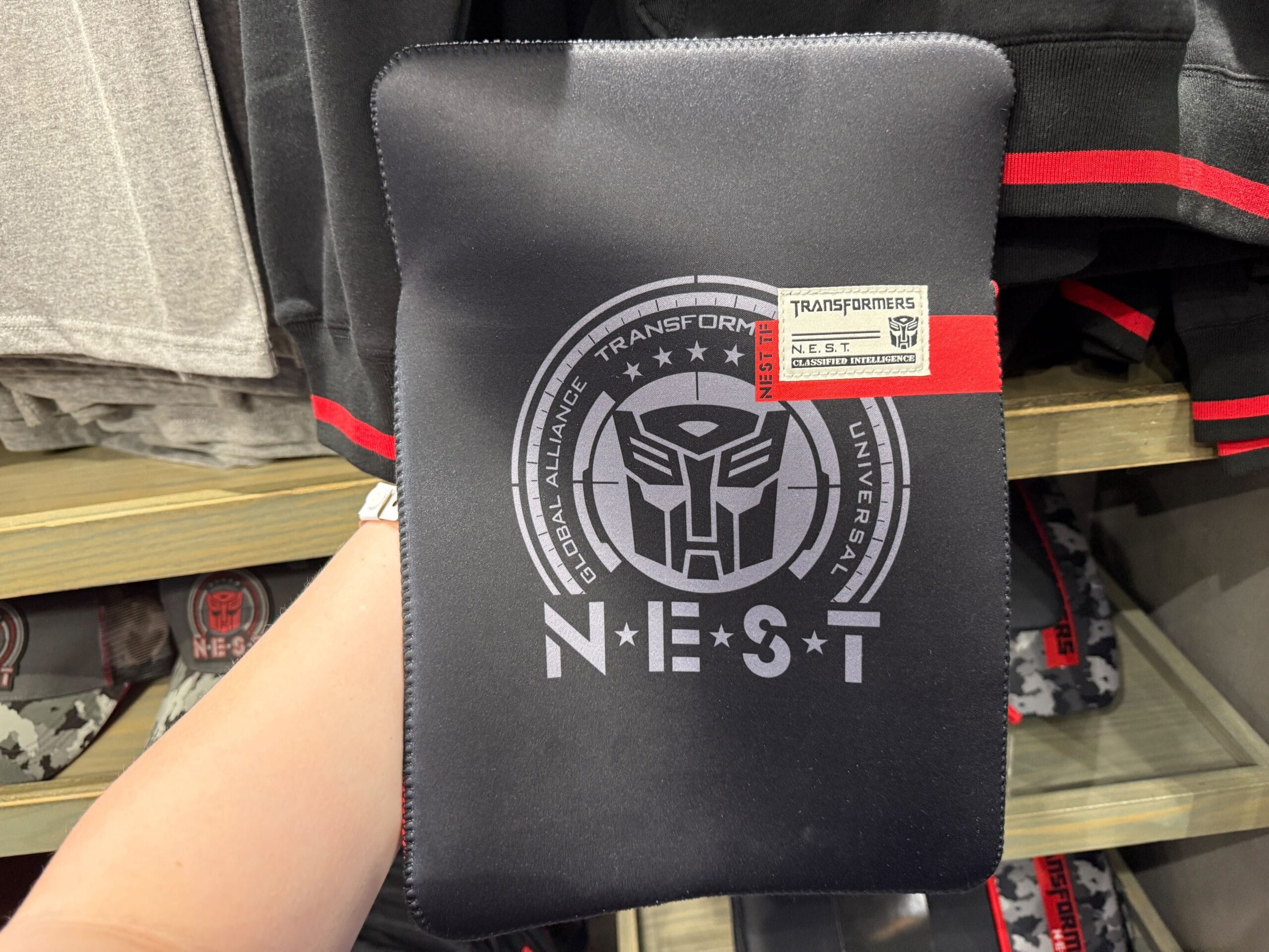 A hand holds a black Transformers N.E.S.T. pouch with a silver logo and label, displayed in a store.