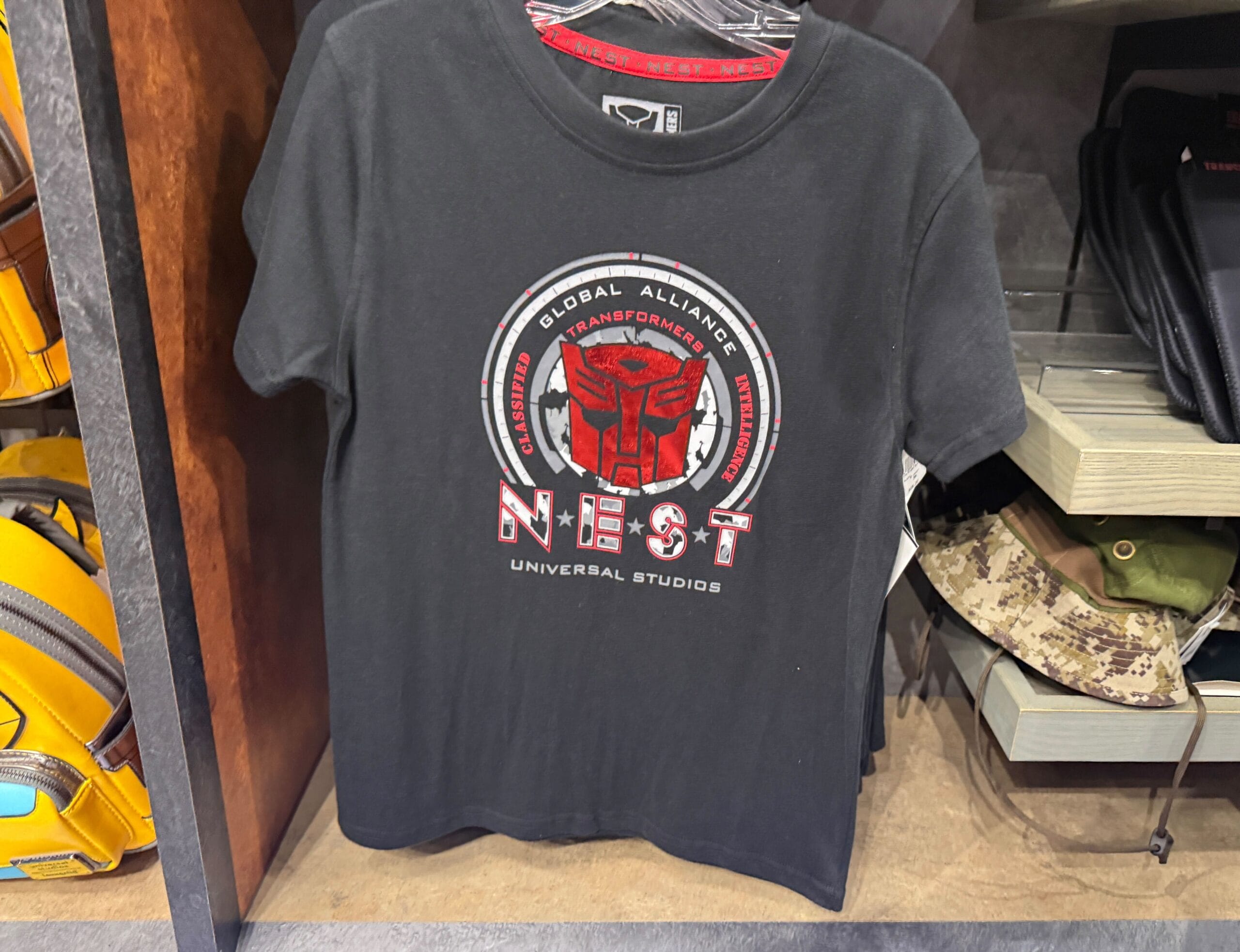 Black T-shirt with a red Transformers logo and “NEST Universal Studios” text displayed on a store rack.