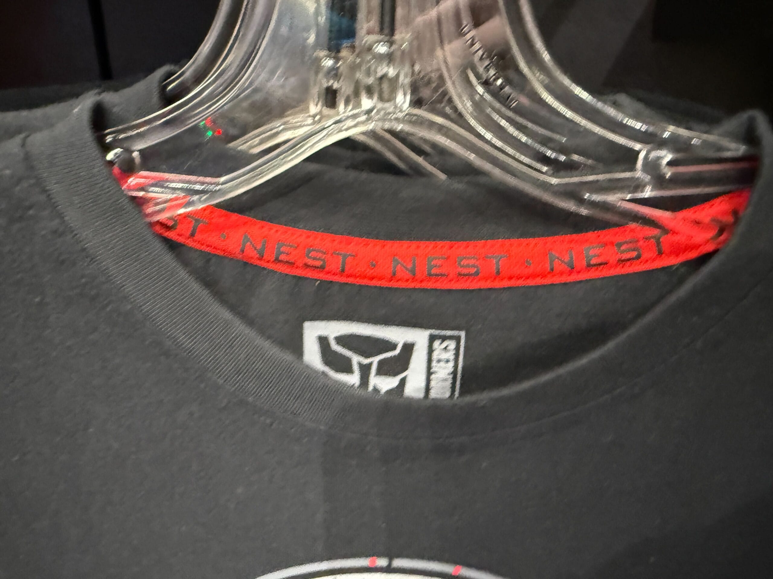 Close-up of a black shirt on a hanger, with a red collar band reading "NEST" and a Transformers Autobot logo tag.