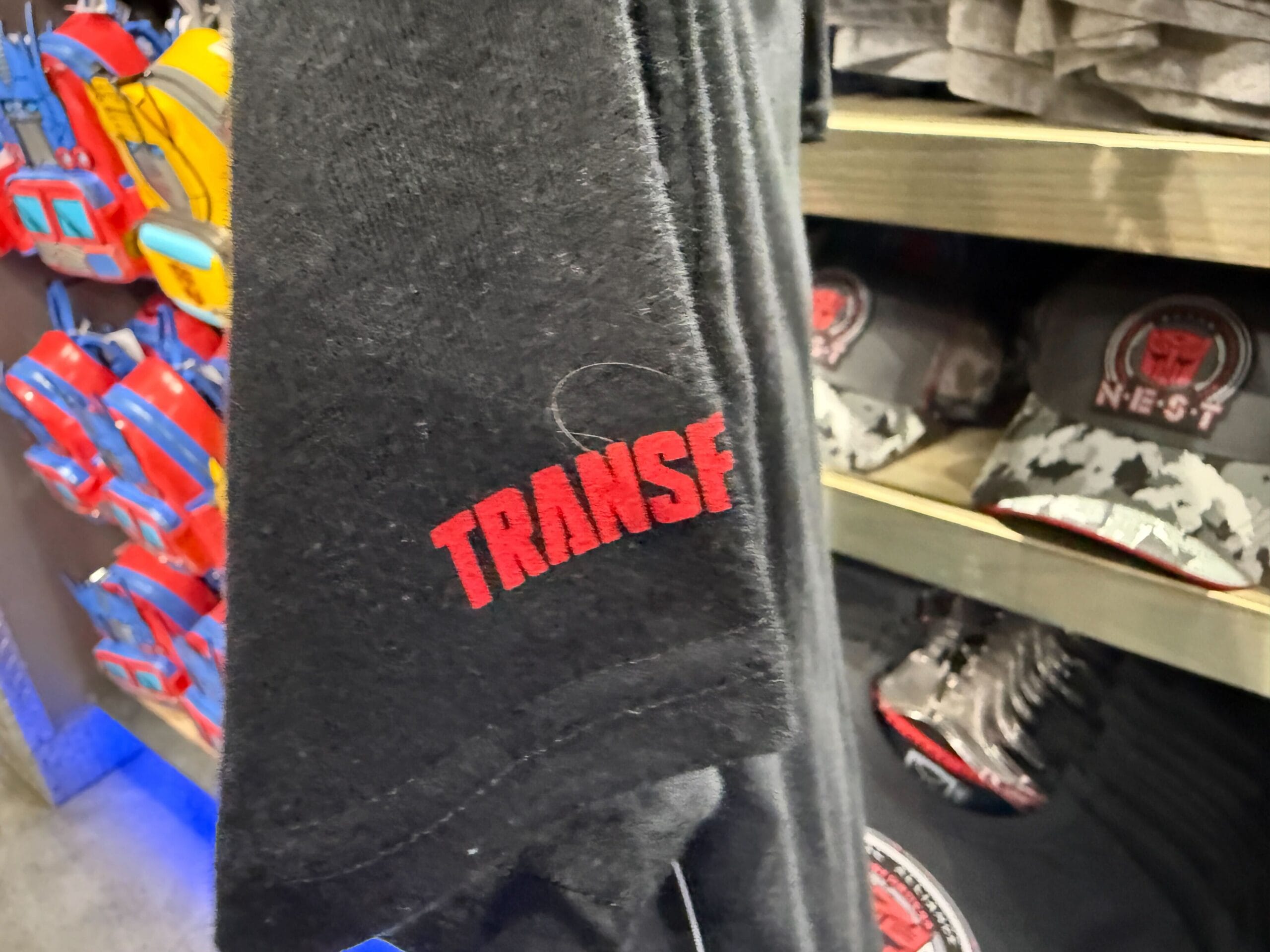 Close-up of a black T-shirt with “TRANSF” in red letters, displayed in a store with themed caps and items in the background.