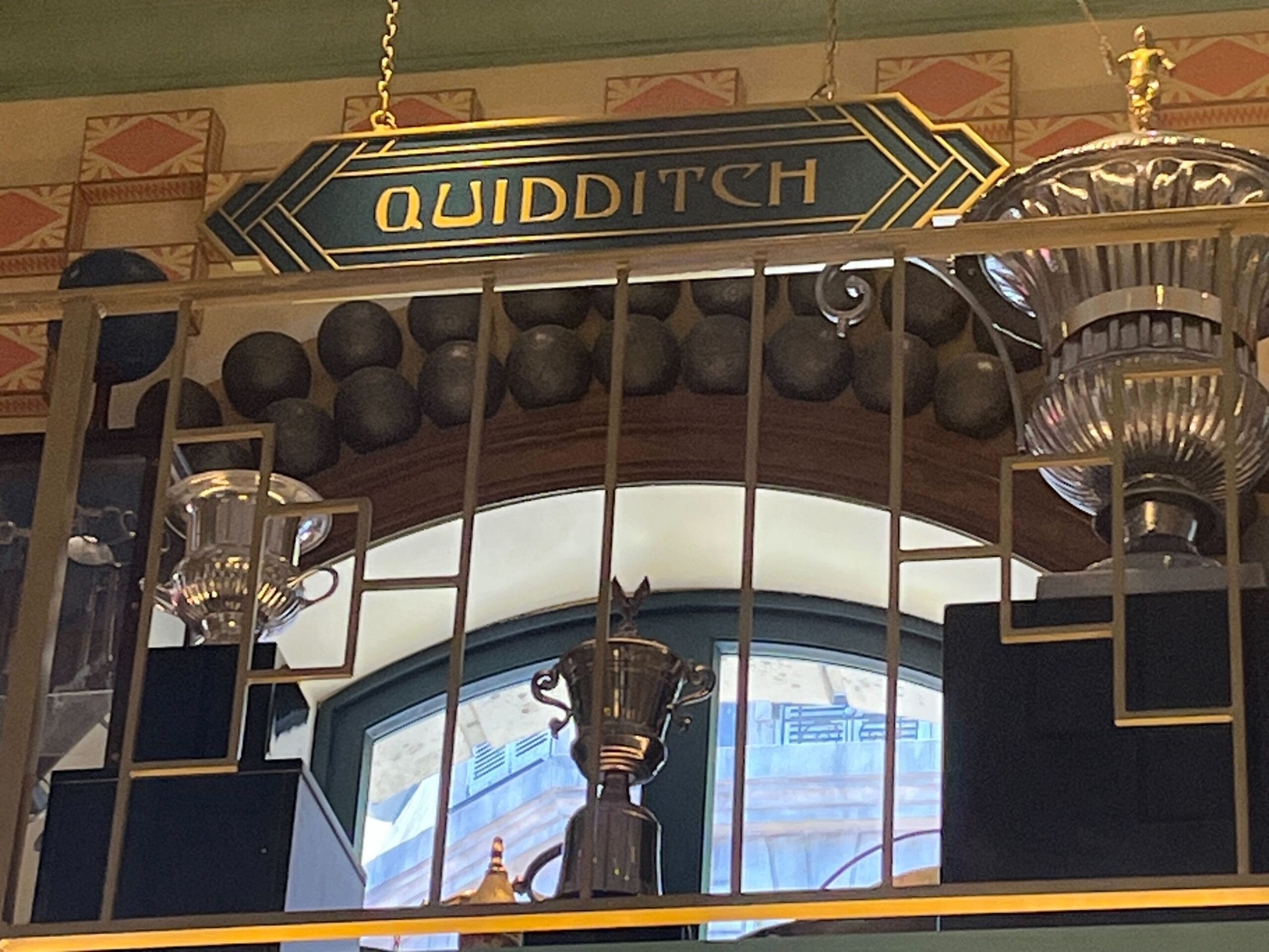 A "Quidditch" sign hangs above trophies and black balls at Les Galeries Mirifiques, on a shelf with a decorative railing.