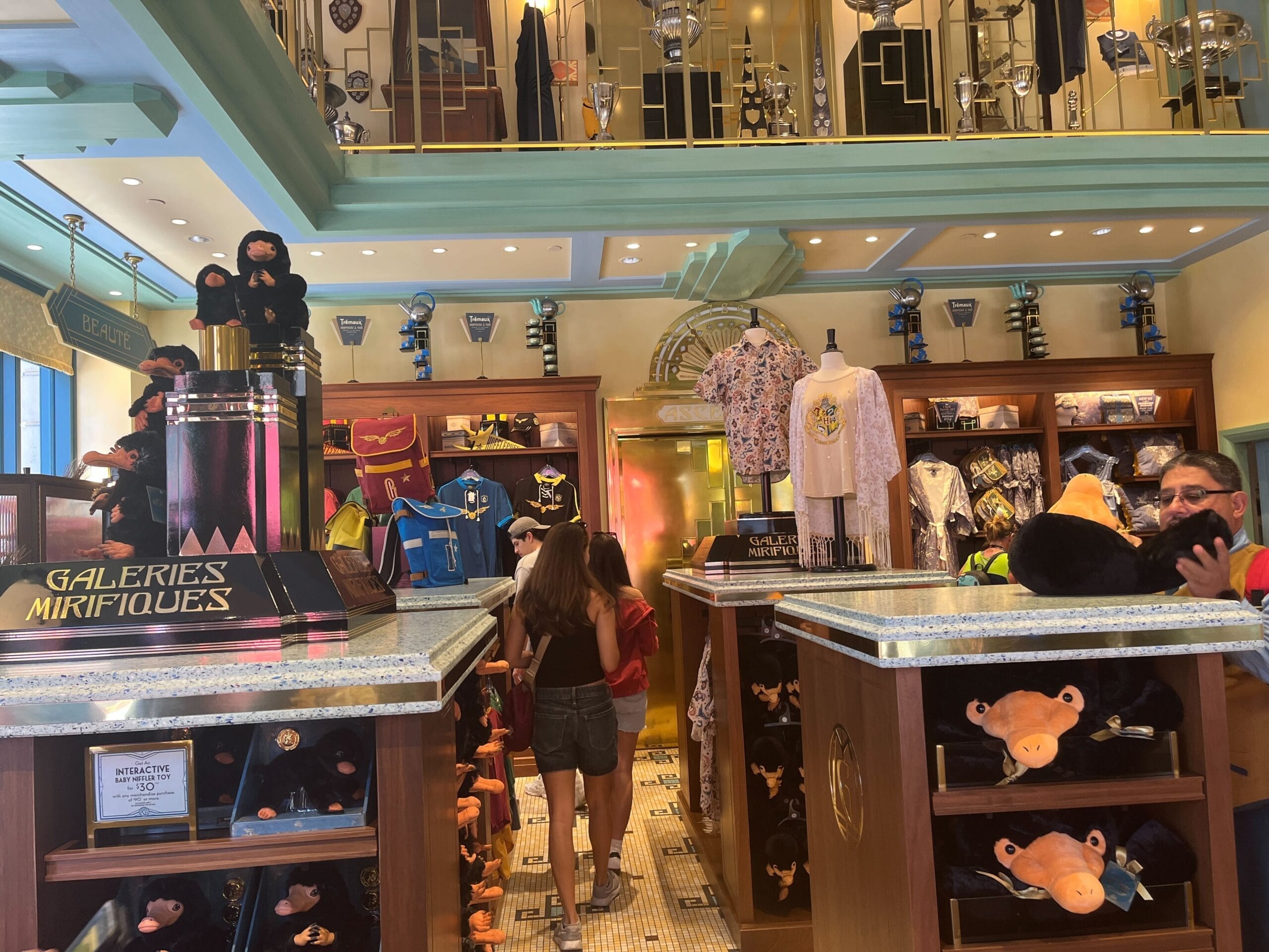 A shop interior at Les Galeries Mirifiques with plush Niffler toys, Harry Potter merchandise, and people browsing the displays.