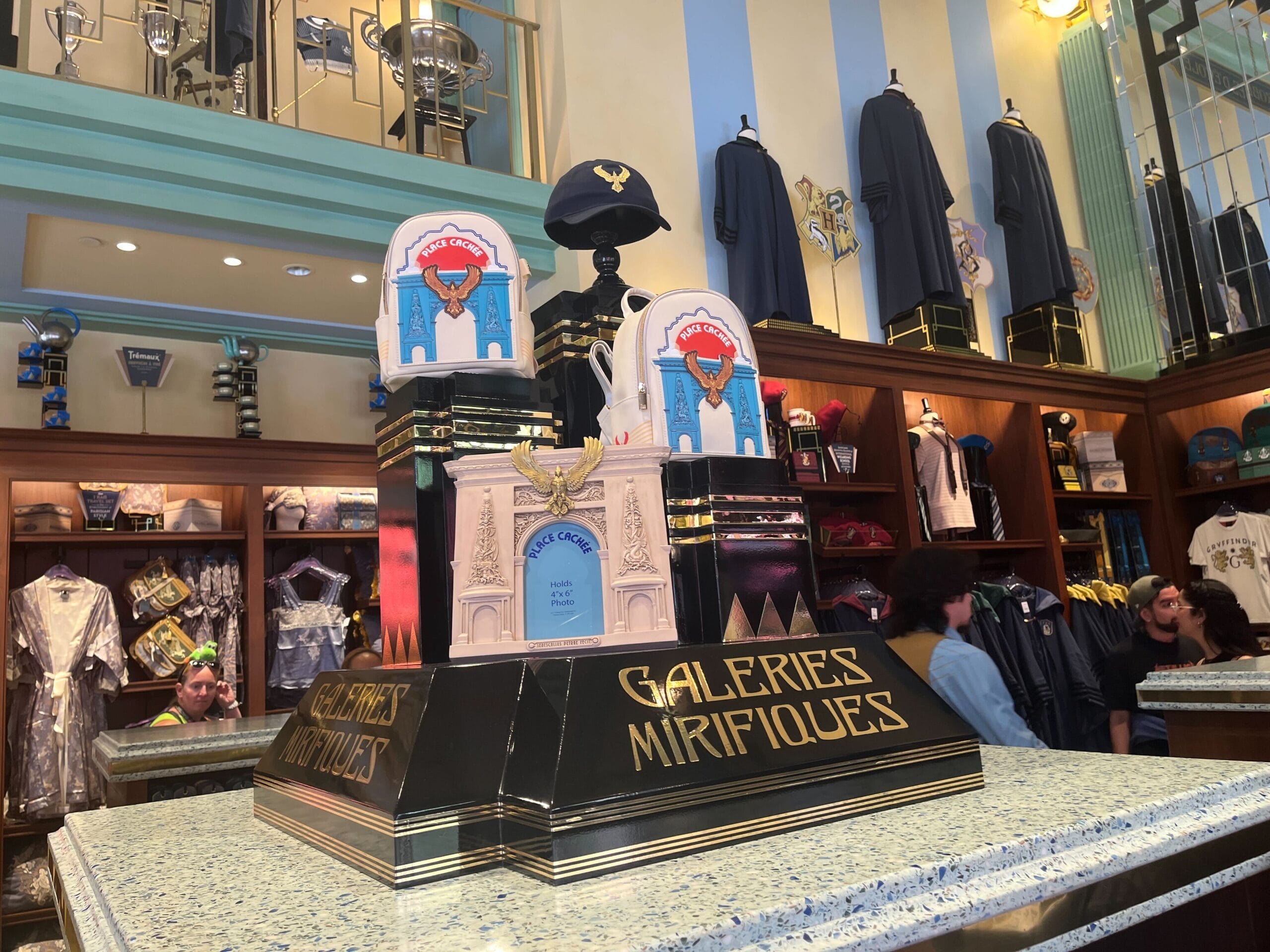 A display labeled "Les Galeries Mirifiques" features themed merchandise in a colorful, decorative store with shoppers browsing.