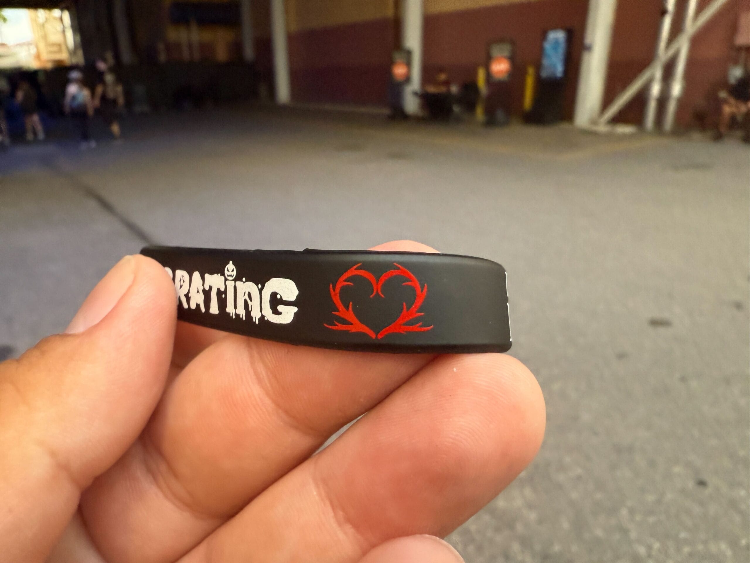A hand holds a black wristband with white text and a red heart-shaped design outdoors.