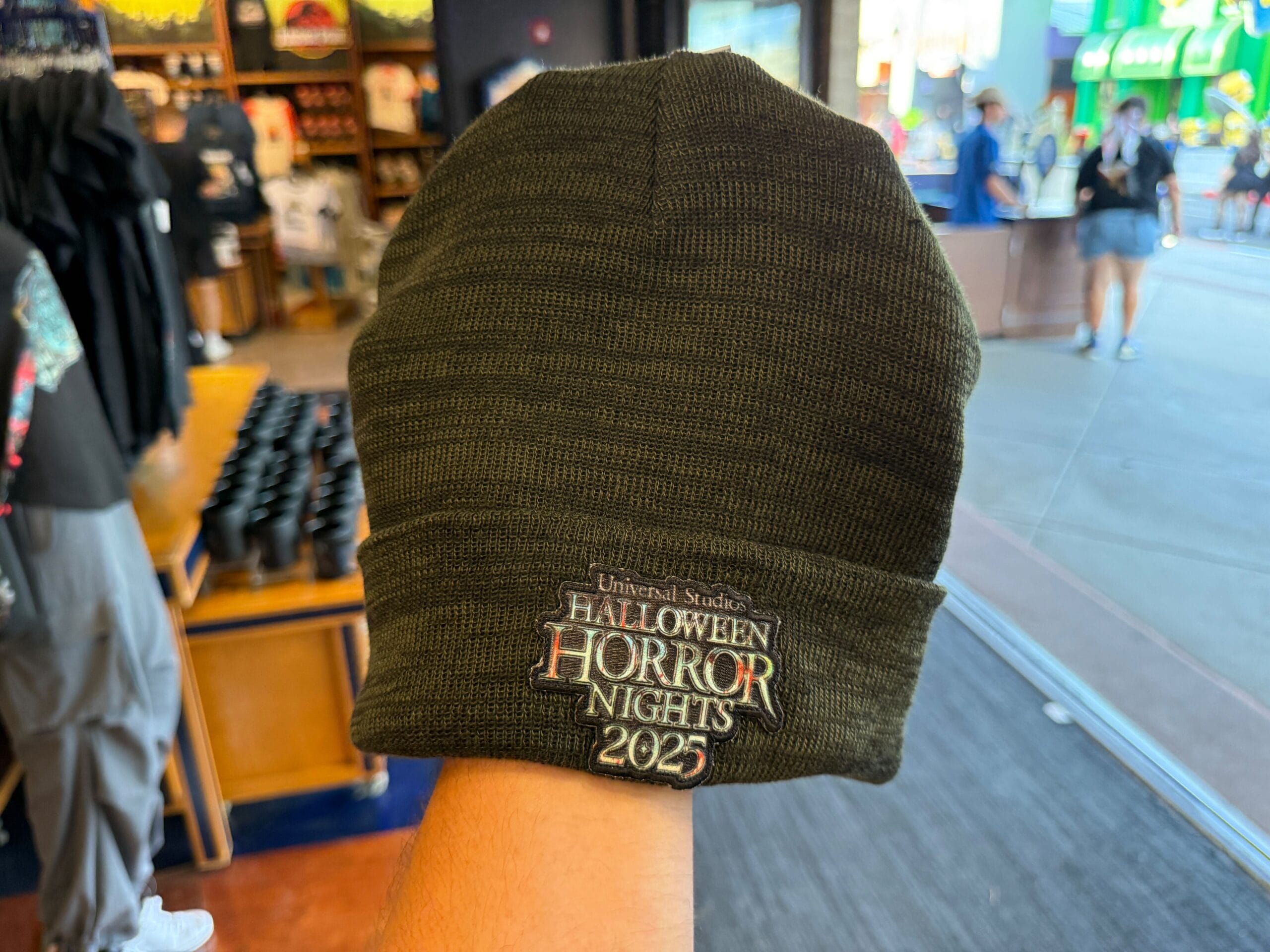A hand holds a green beanie with "Halloween Horror Nights 2025" embroidered on the front in a store.