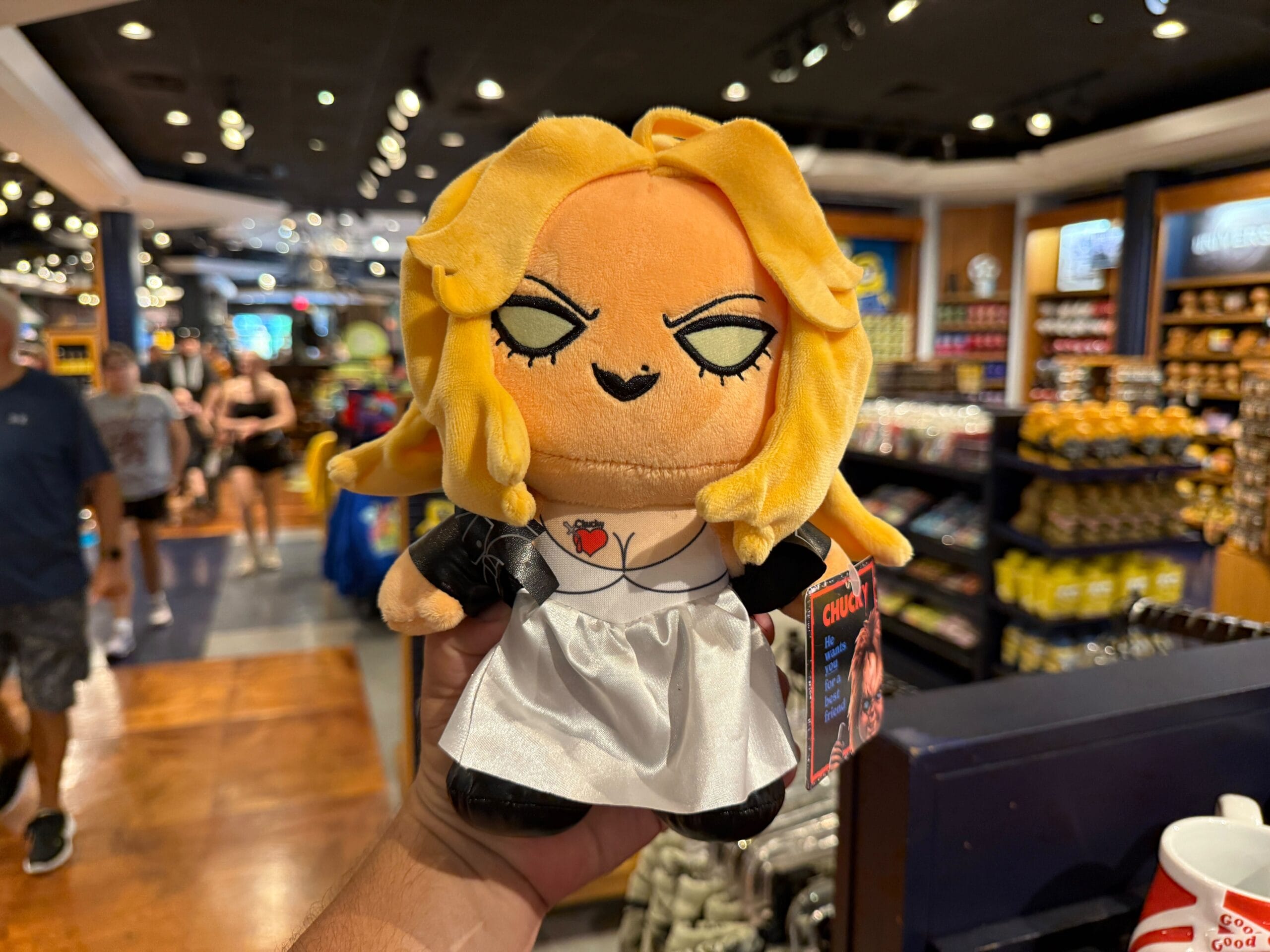 A hand holds a plush doll with blonde hair and a white dress in a busy gift shop.
