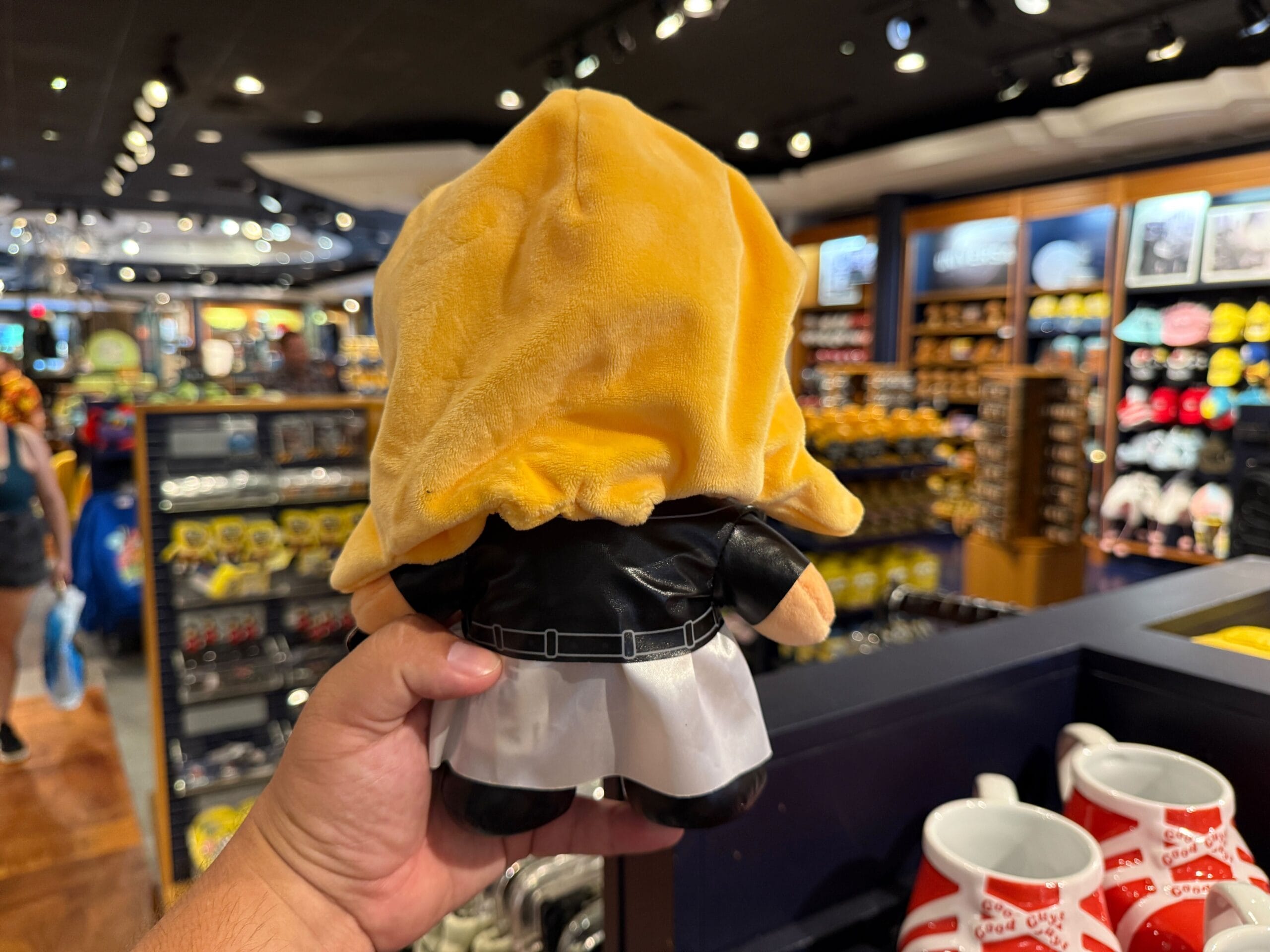 A hand holds a plush toy with a yellow hood and white skirt in a store filled with various merchandise.