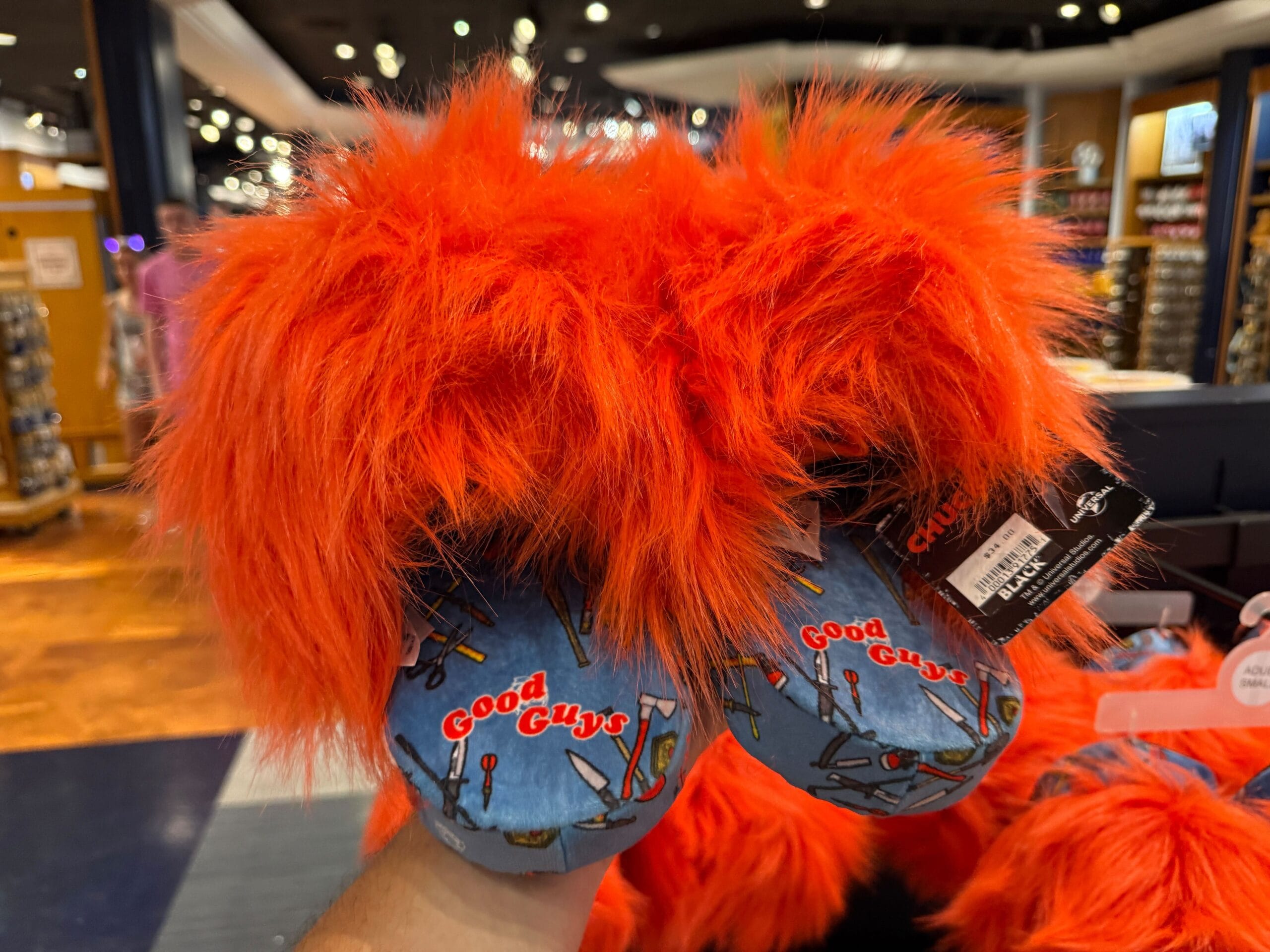 A hand holds fuzzy orange slippers with “Good Guys” and weapon patterns on blue soles in a store.
