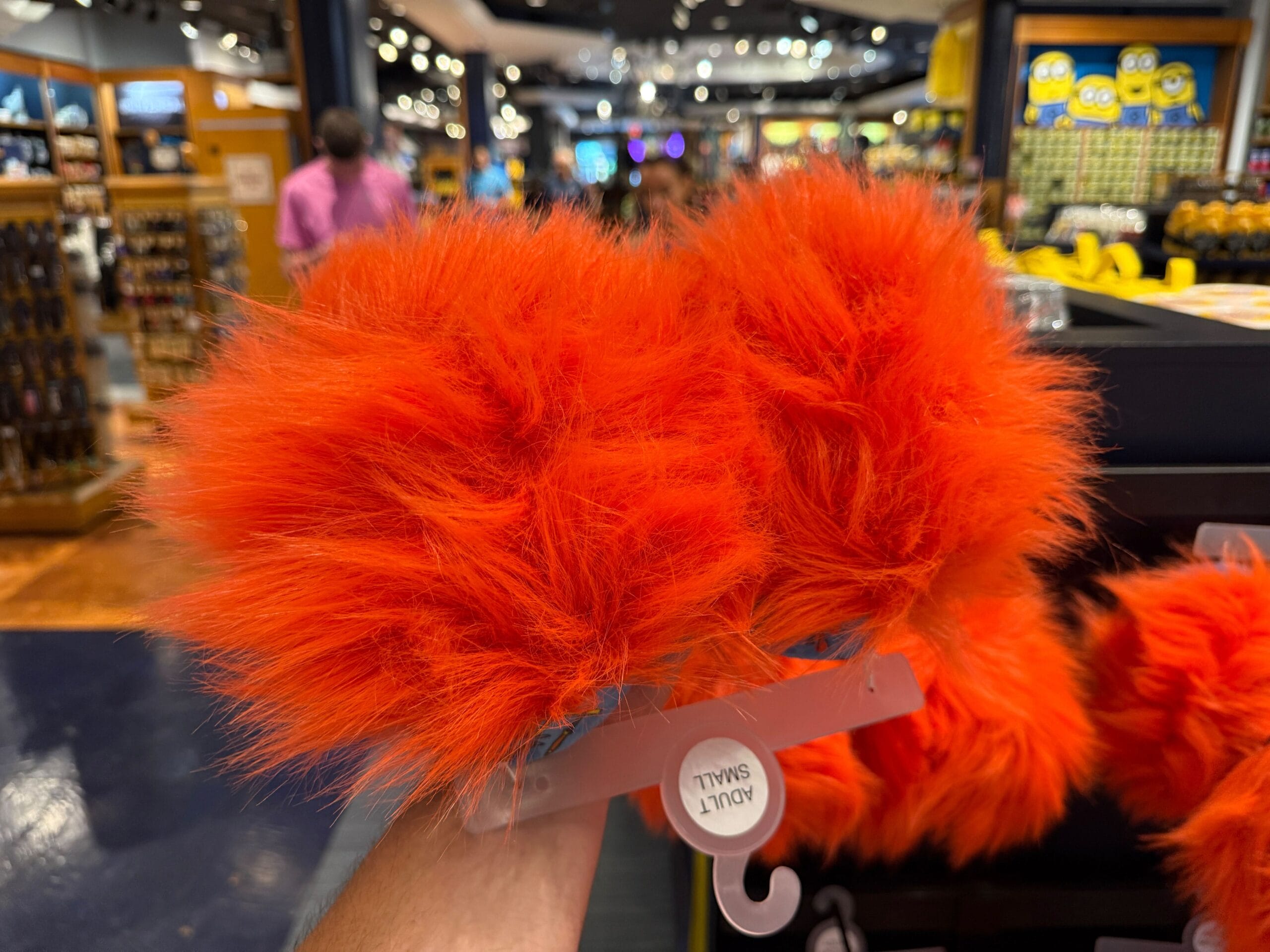 A hand holds a pair of bright orange furry slippers labeled "ADULT SMALL" in a store with colorful displays.
