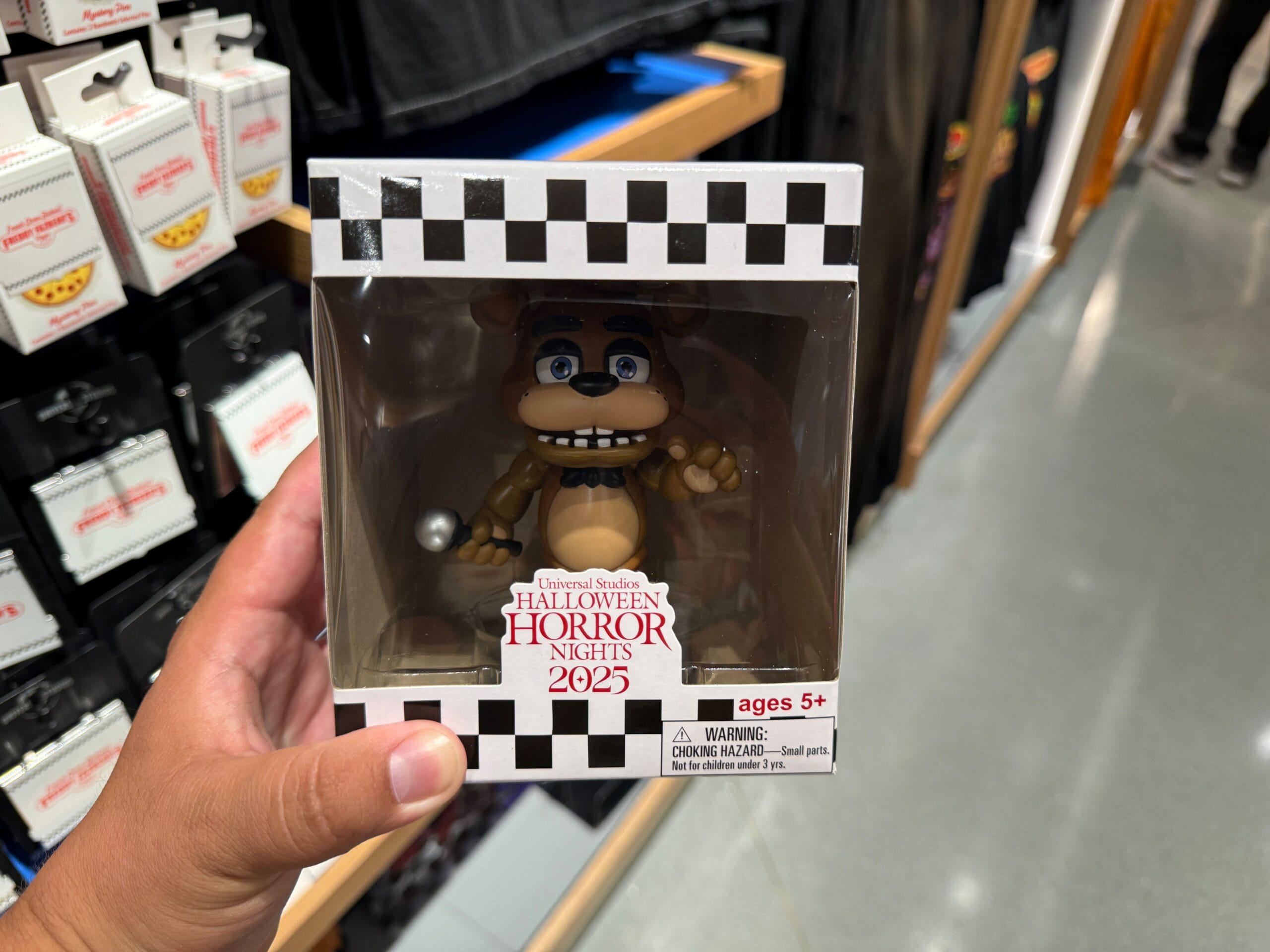 Hand holding a "Halloween Horror Nights 2025" Freddy Fazbear figure box in a store aisle.
