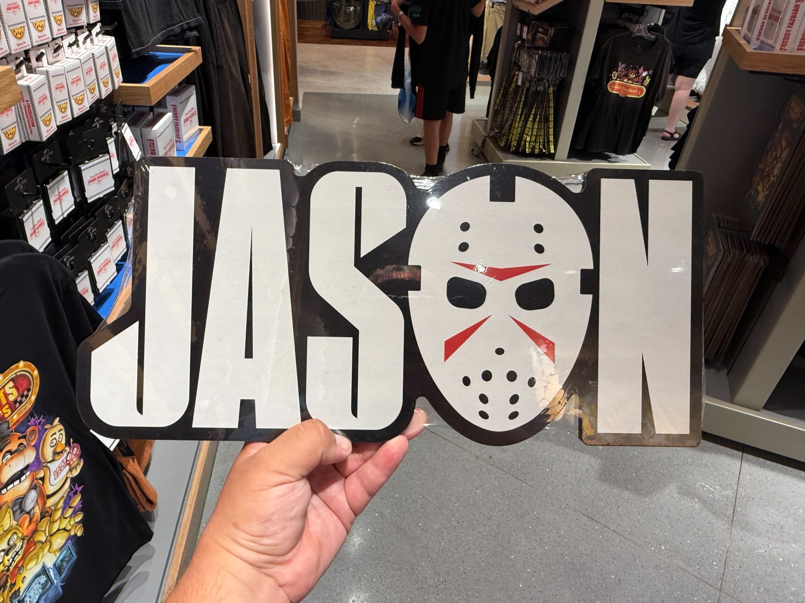 A hand holds a "JASON" sign with a hockey mask forming the letter "O" in a store.