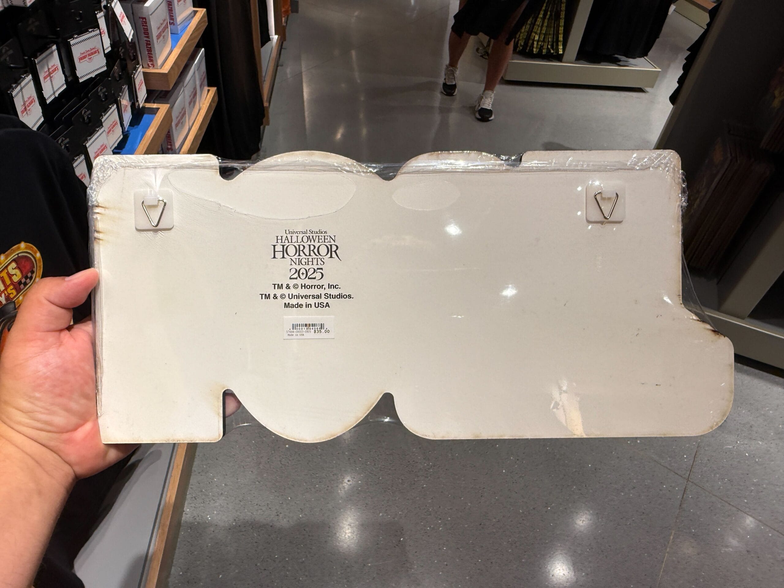 A hand holds a cardboard sign labeled "Halloween Horror Nights 2023" in a store with shiny gray flooring.