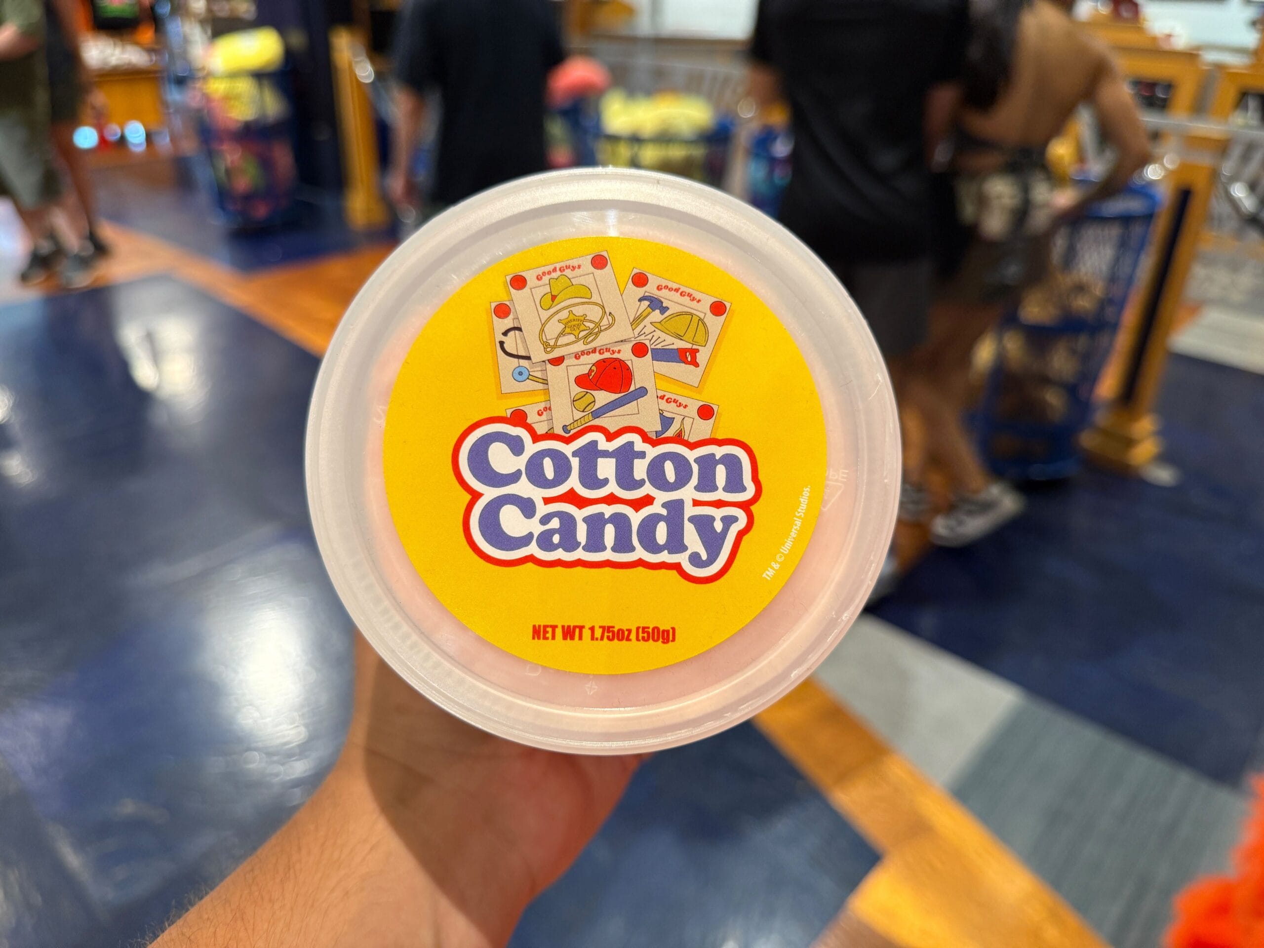 A hand holds a round container labeled "Cotton Candy" with a yellow lid in a colorful store setting.