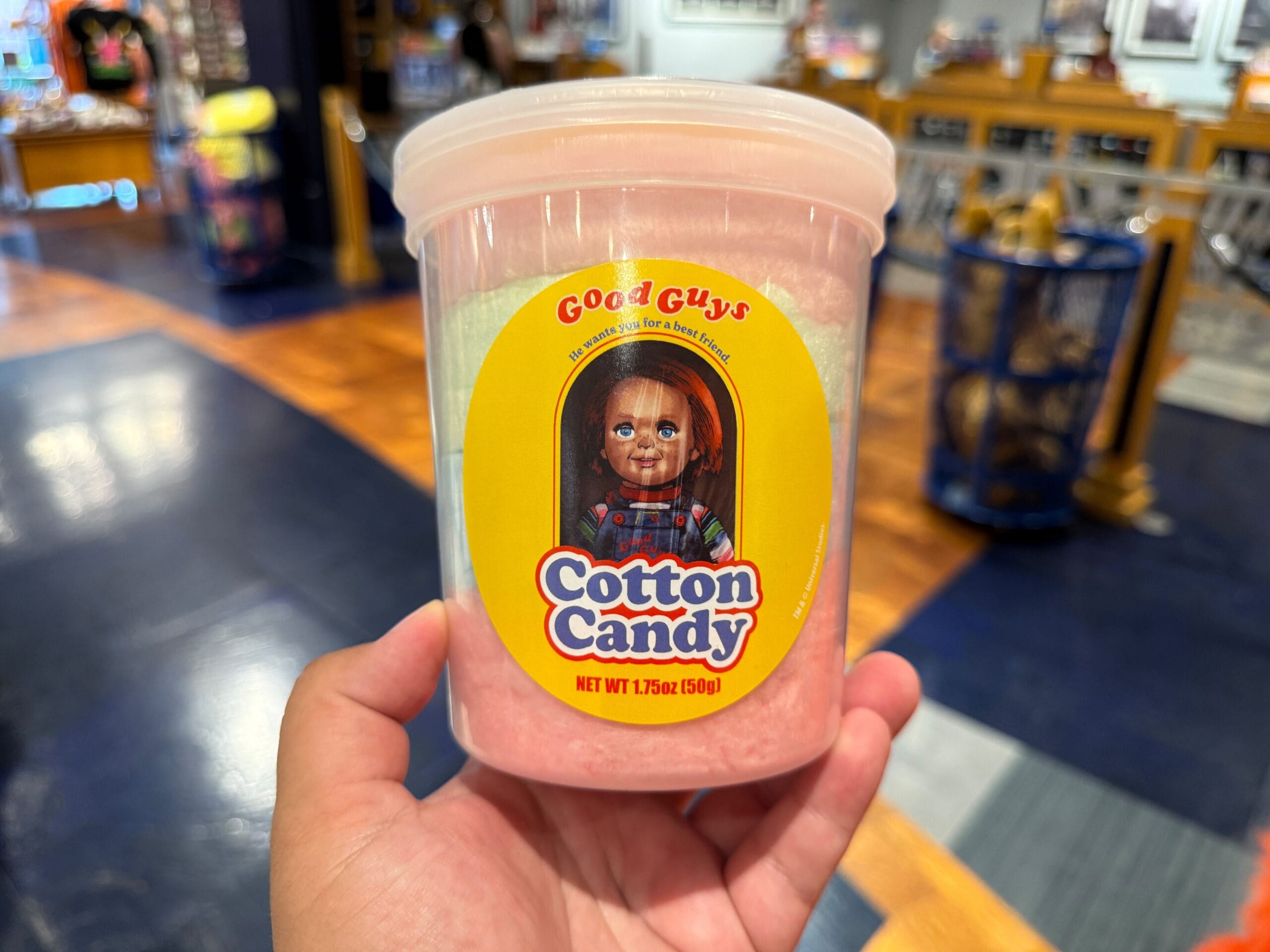 A hand holds a tub of pink cotton candy featuring a "Good Guys" Chucky label in a store.