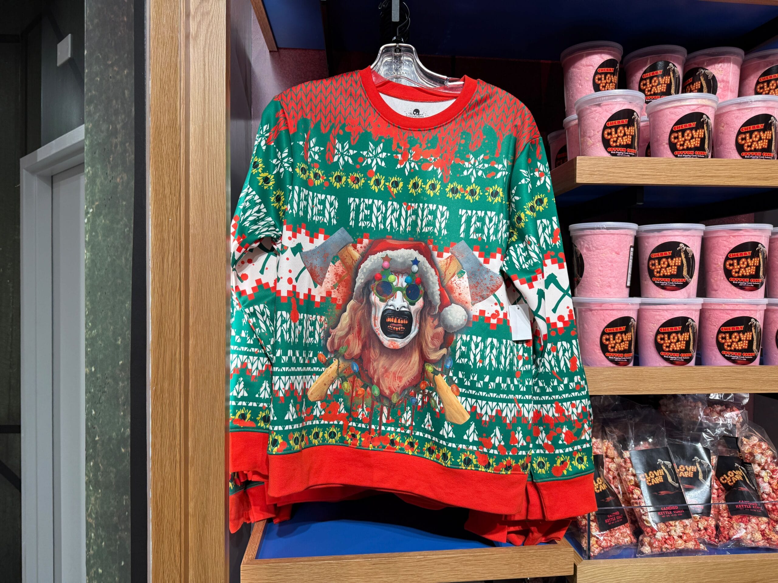 A festive sweater with a Krampus design hangs on a rack near shelves of pink Clown Cafe popcorn tubs.