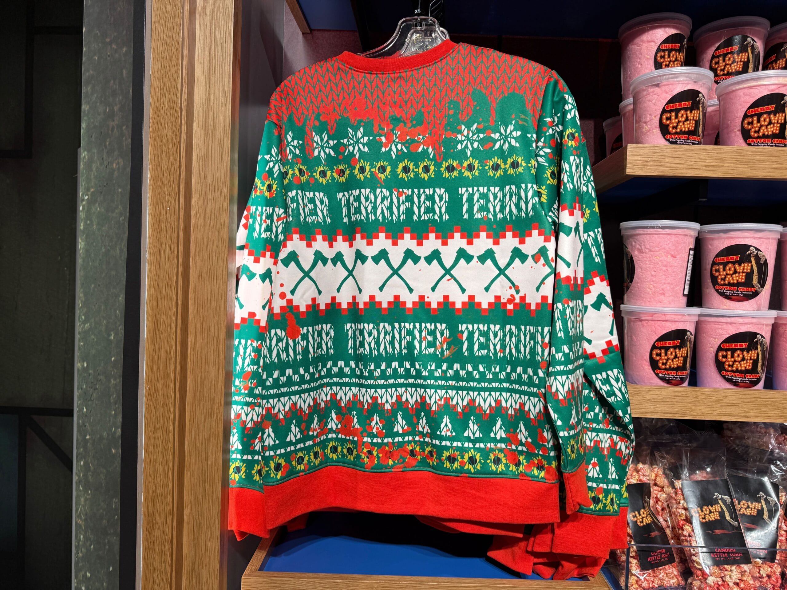 A festive "TERRIFIER" holiday sweater with red, green, and white patterns hangs on display next to shelves of candy.