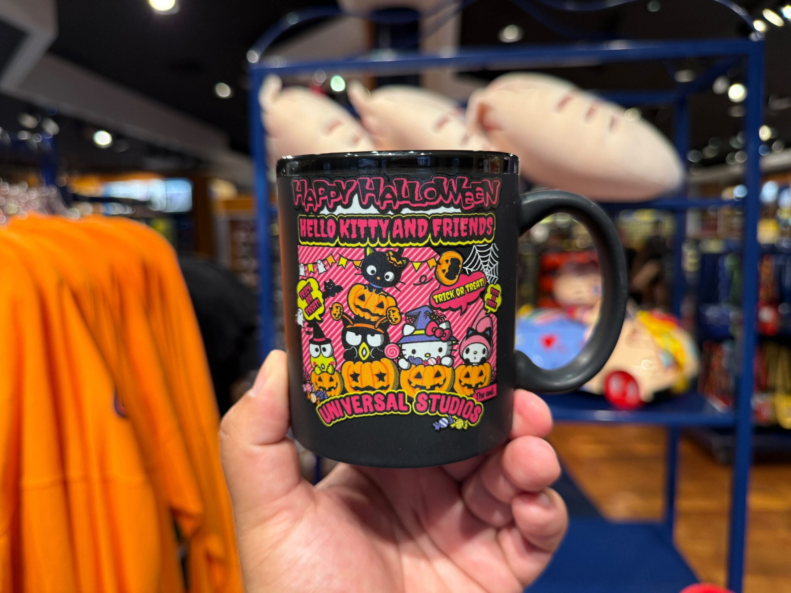 A hand holds a black mug with Hello Kitty and friends in Halloween costumes, in a store with colorful displays.