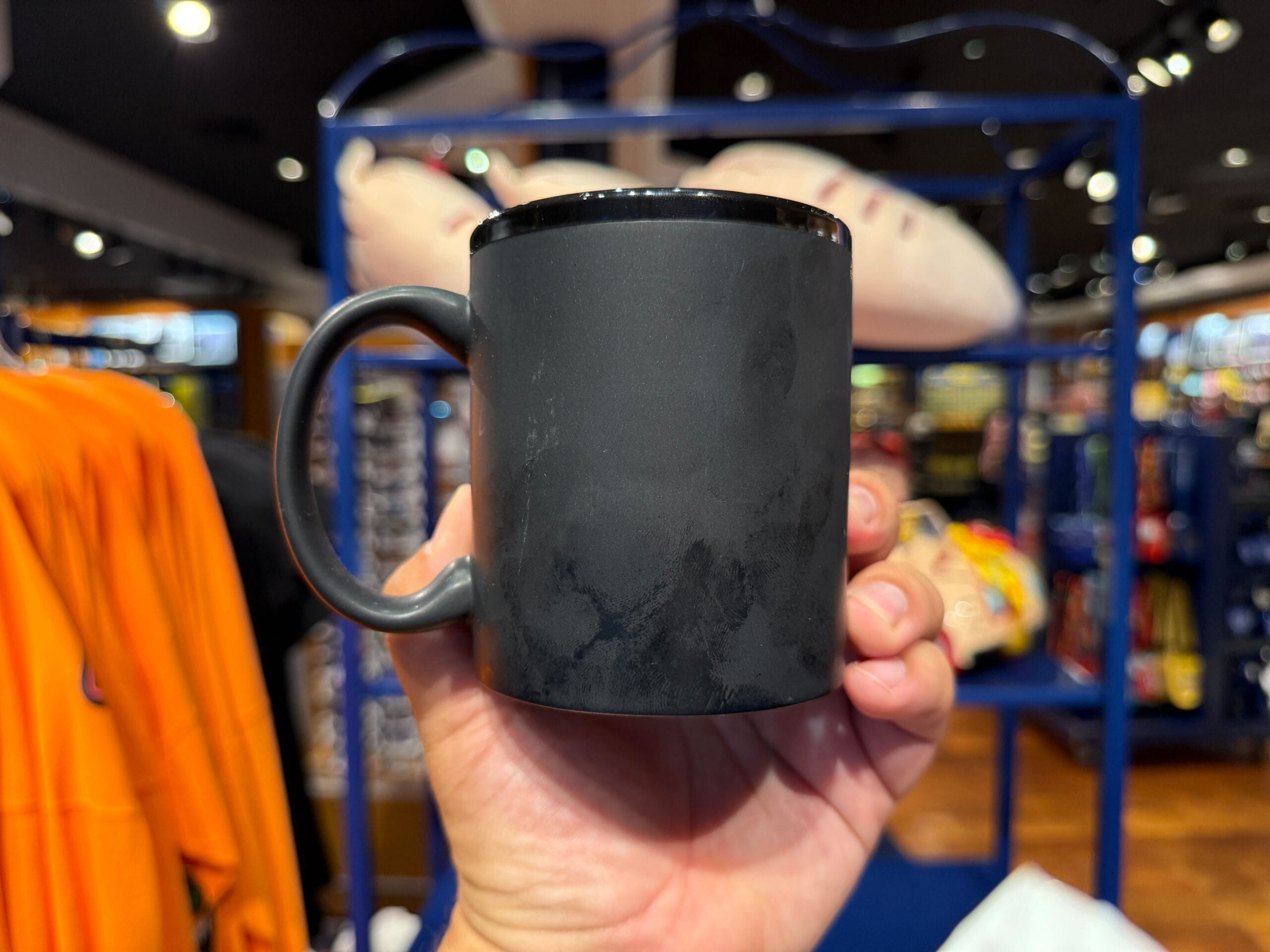 A hand holds a plain black mug inside a brightly lit store with colorful items in the background.