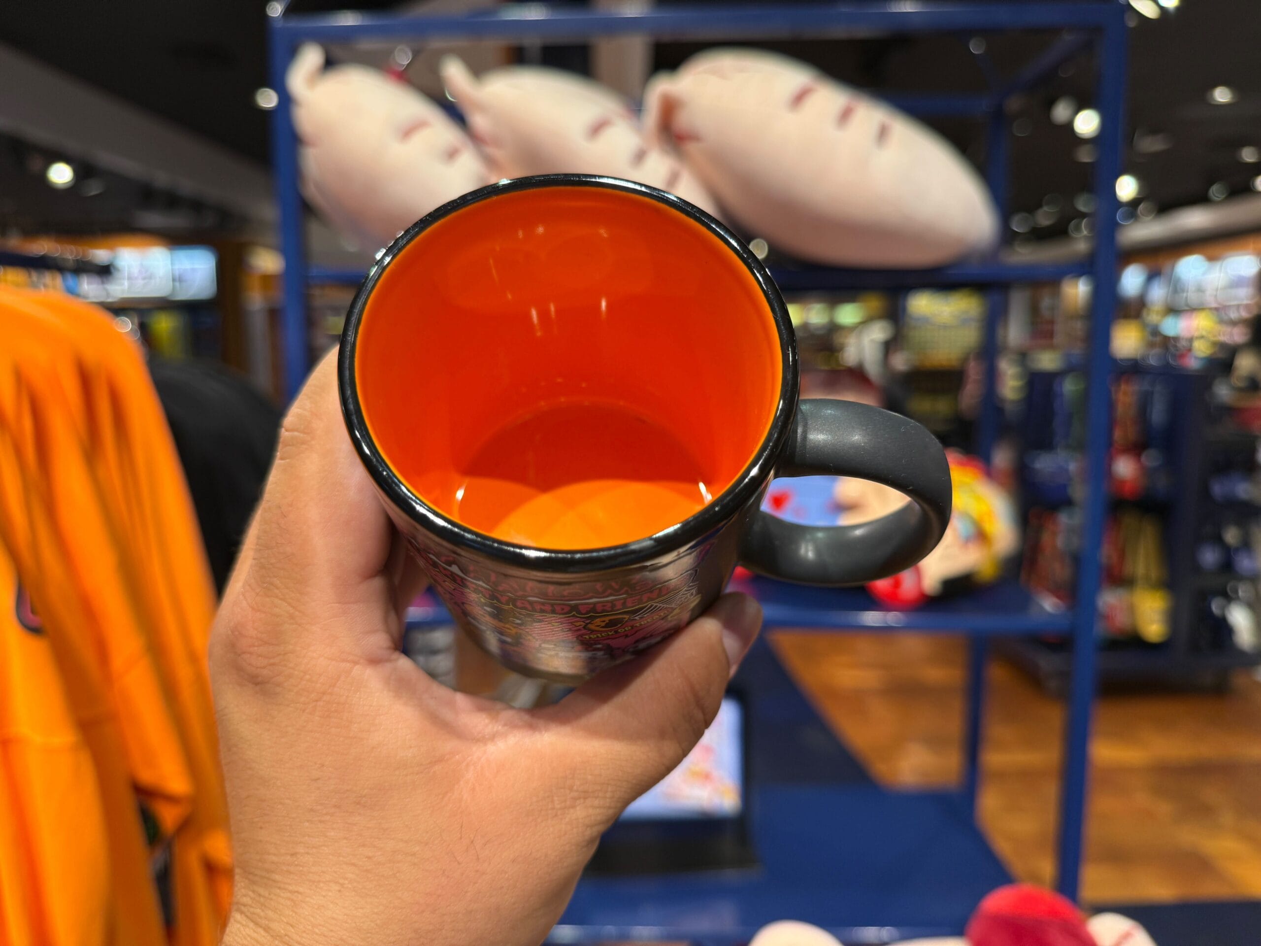 A hand holds a black mug with an orange interior inside a store with plush toys and merchandise in the background.
