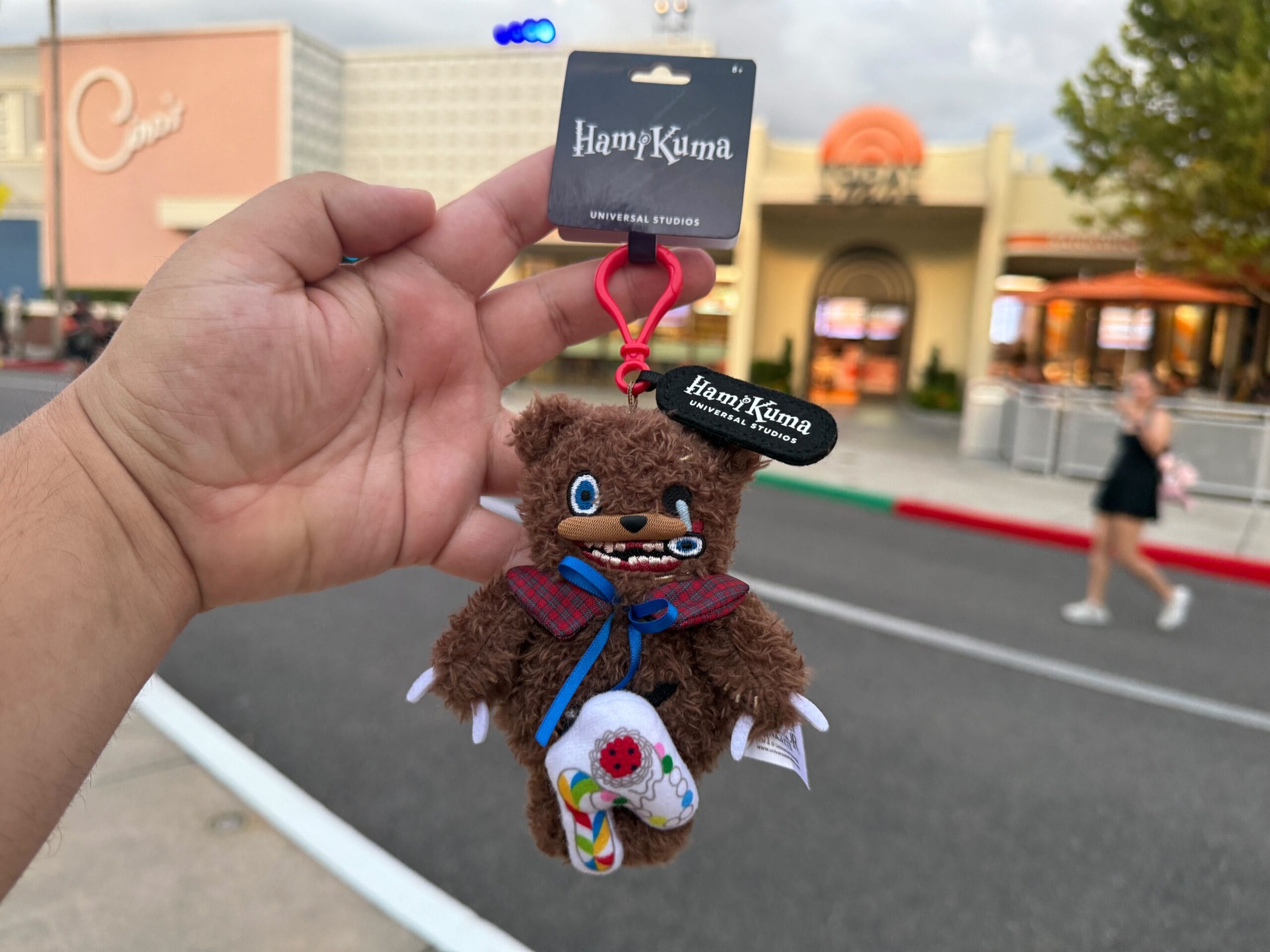 A hand holds a HamKuma Universal Studios keychain featuring a plush bear with stitches and a red clip, outdoors.