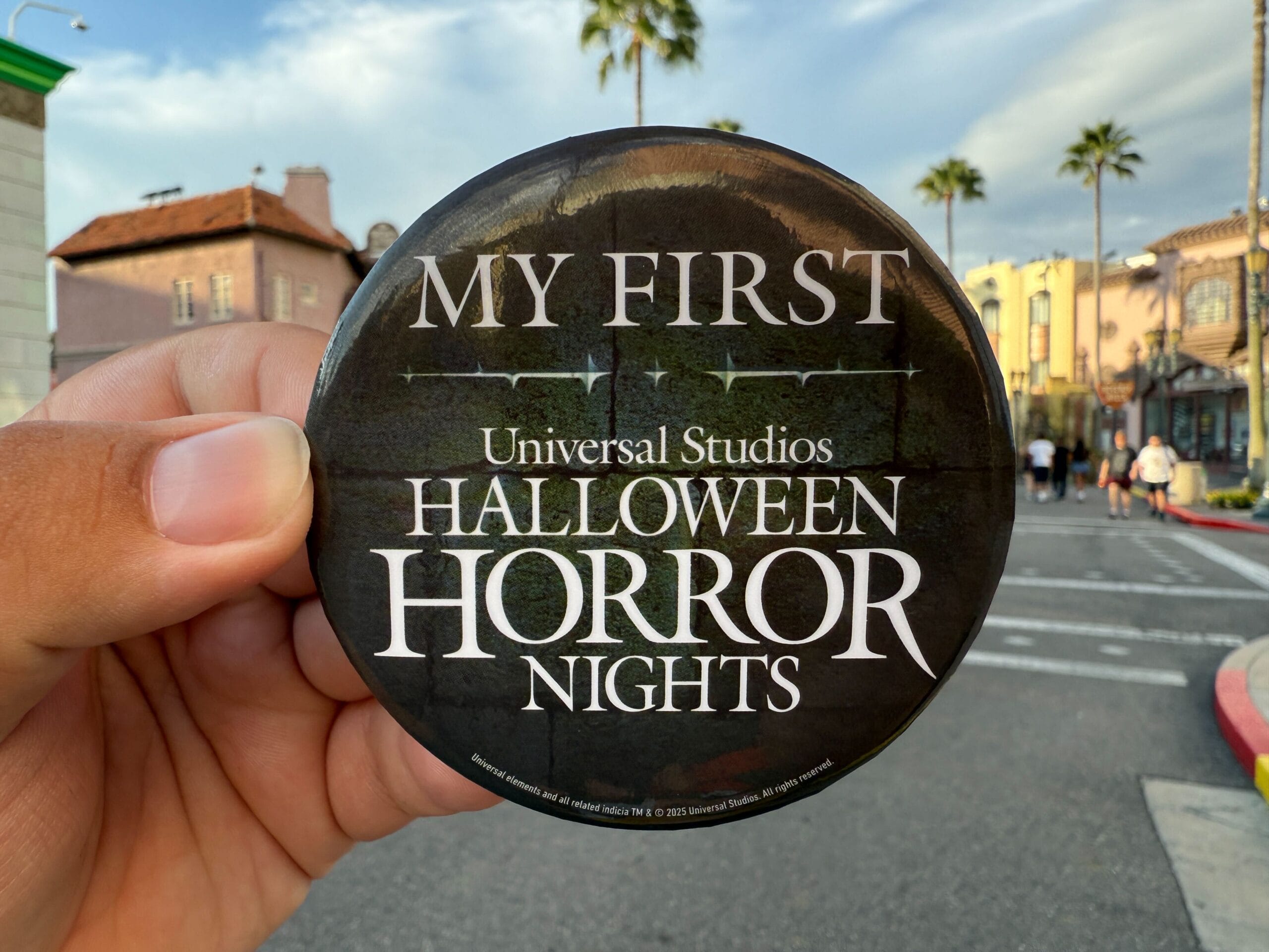 A hand holds a "My First Universal Studios Halloween Horror Nights" button in an outdoor setting.