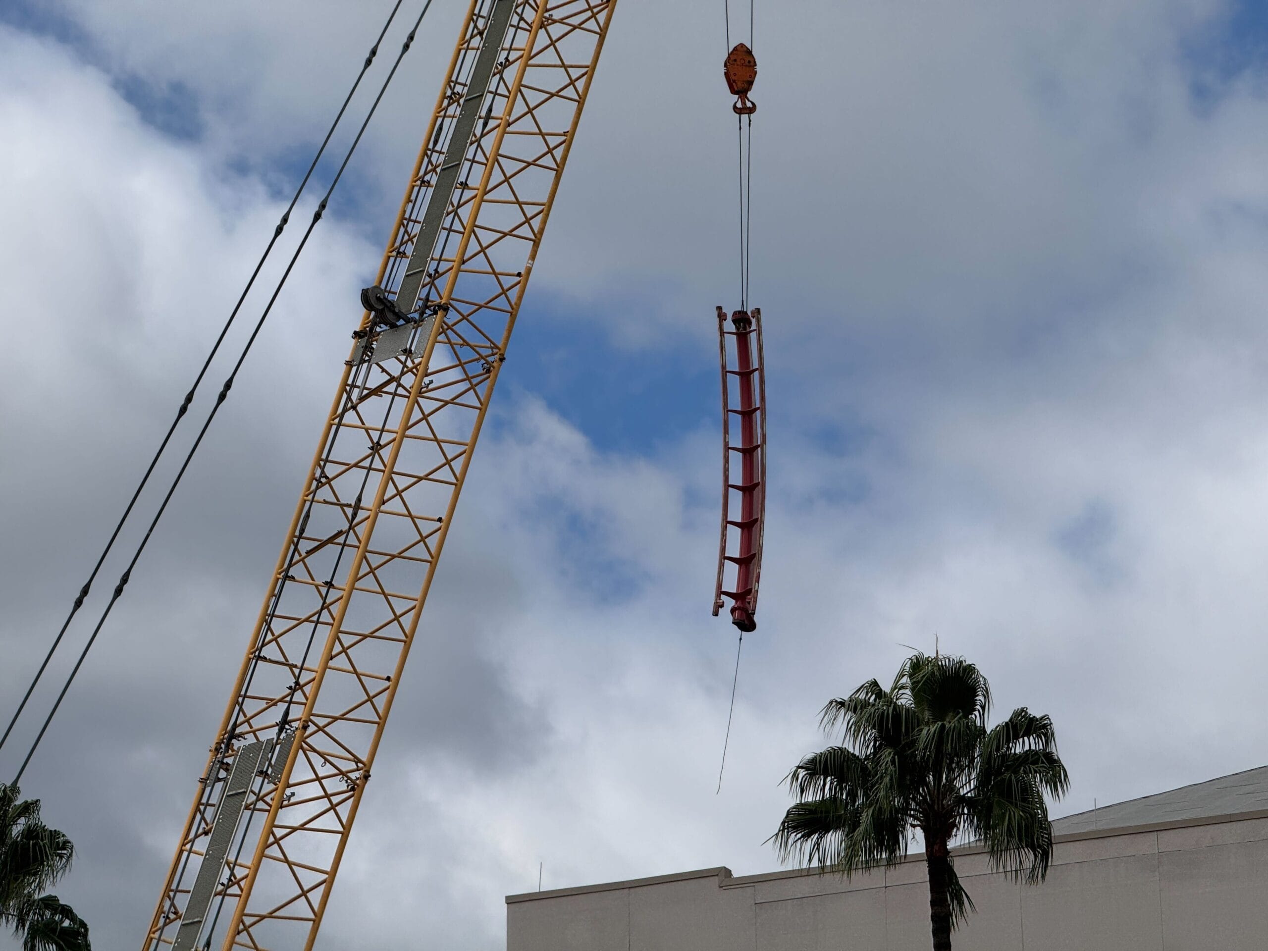 A crane lifts a curved red steel beam in a Rip Ride Rockit demo above a building with palm trees and cloudy skies.