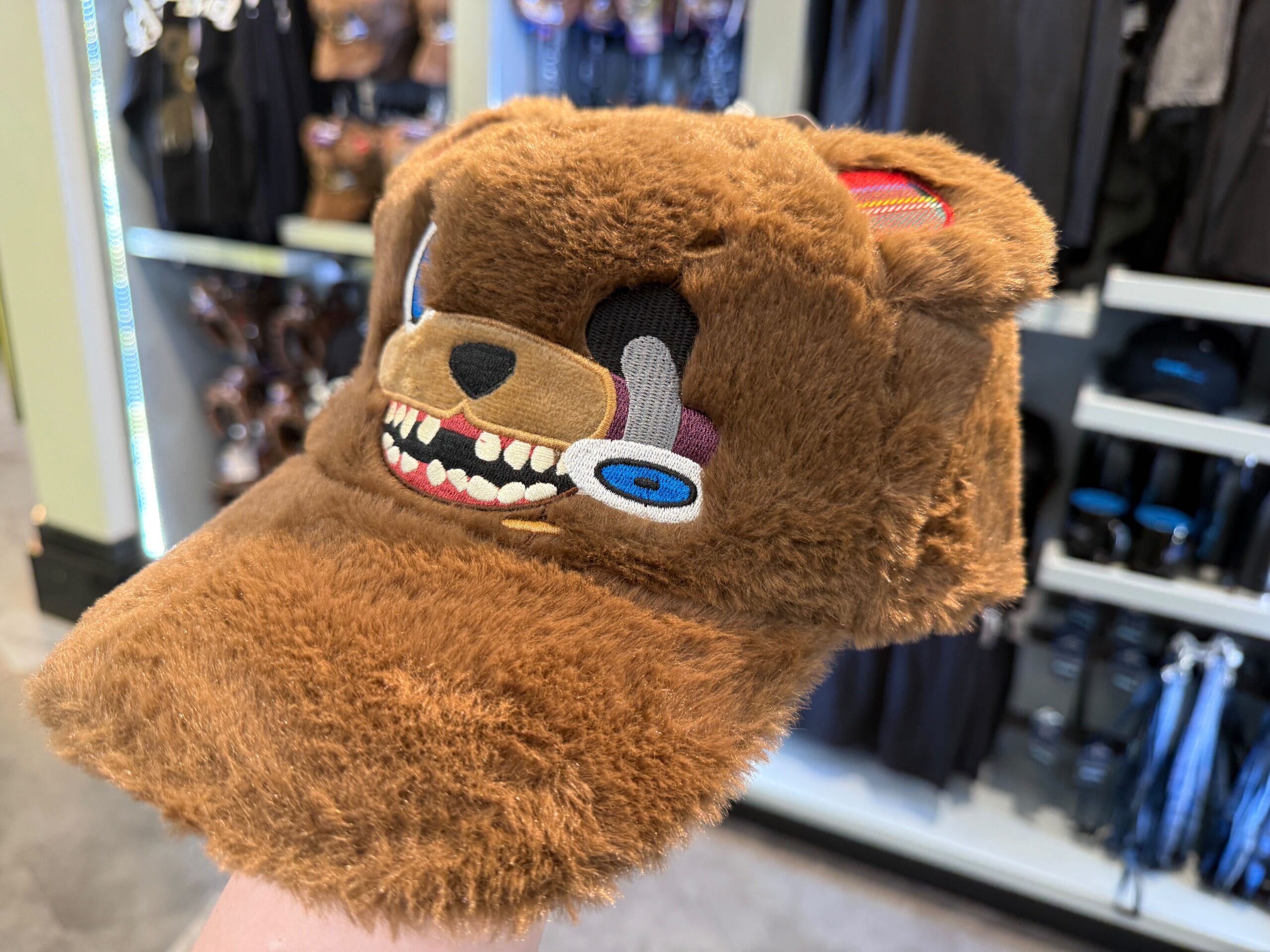 A person holds a fuzzy brown HamiKuma Merchandise cap styled like a cartoonish bear with sharp teeth and blue eyes.