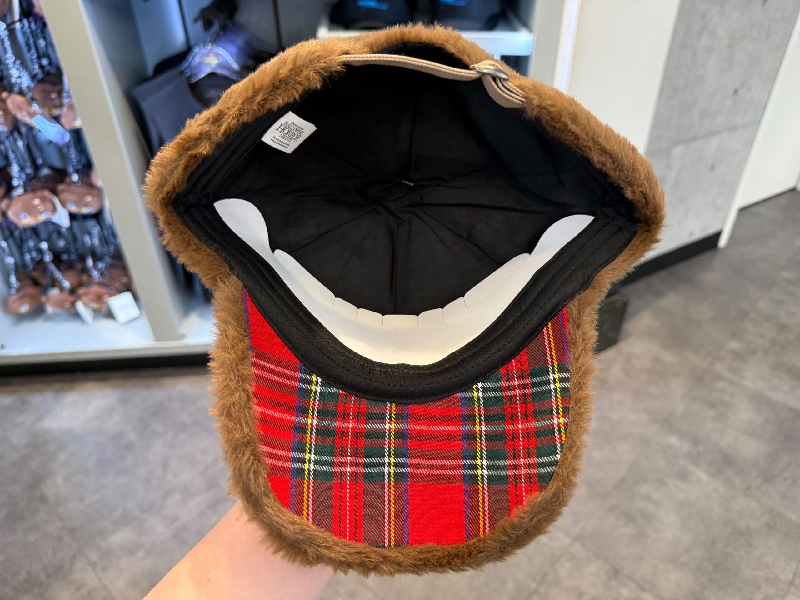 A hand holds a HamiKuma Merchandise furry hat with a red plaid brim and white fake teeth inside the front.