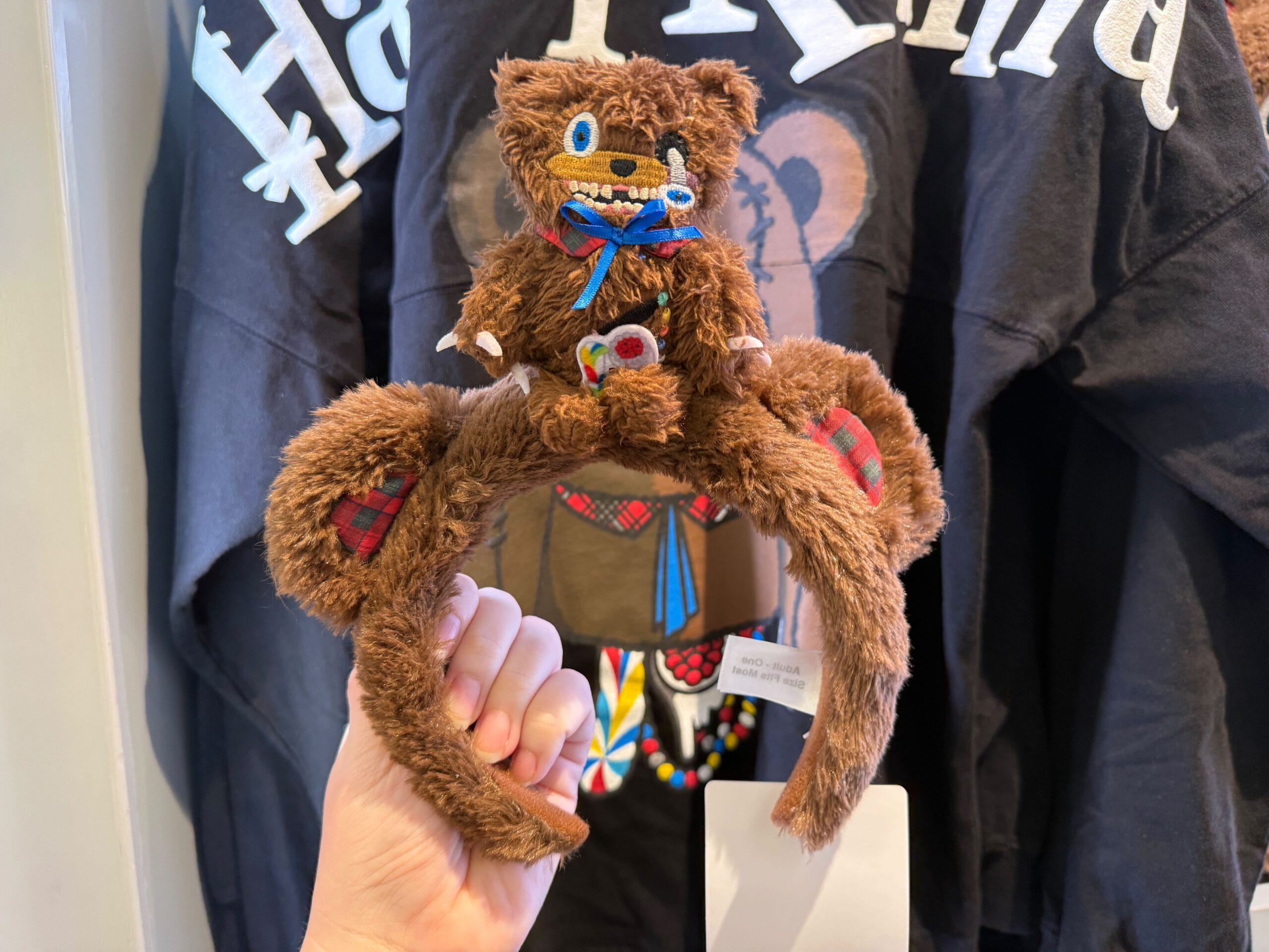 A hand displays a HamiKuma Merchandise headband with a plush bear and plaid, in front of black shirts.
