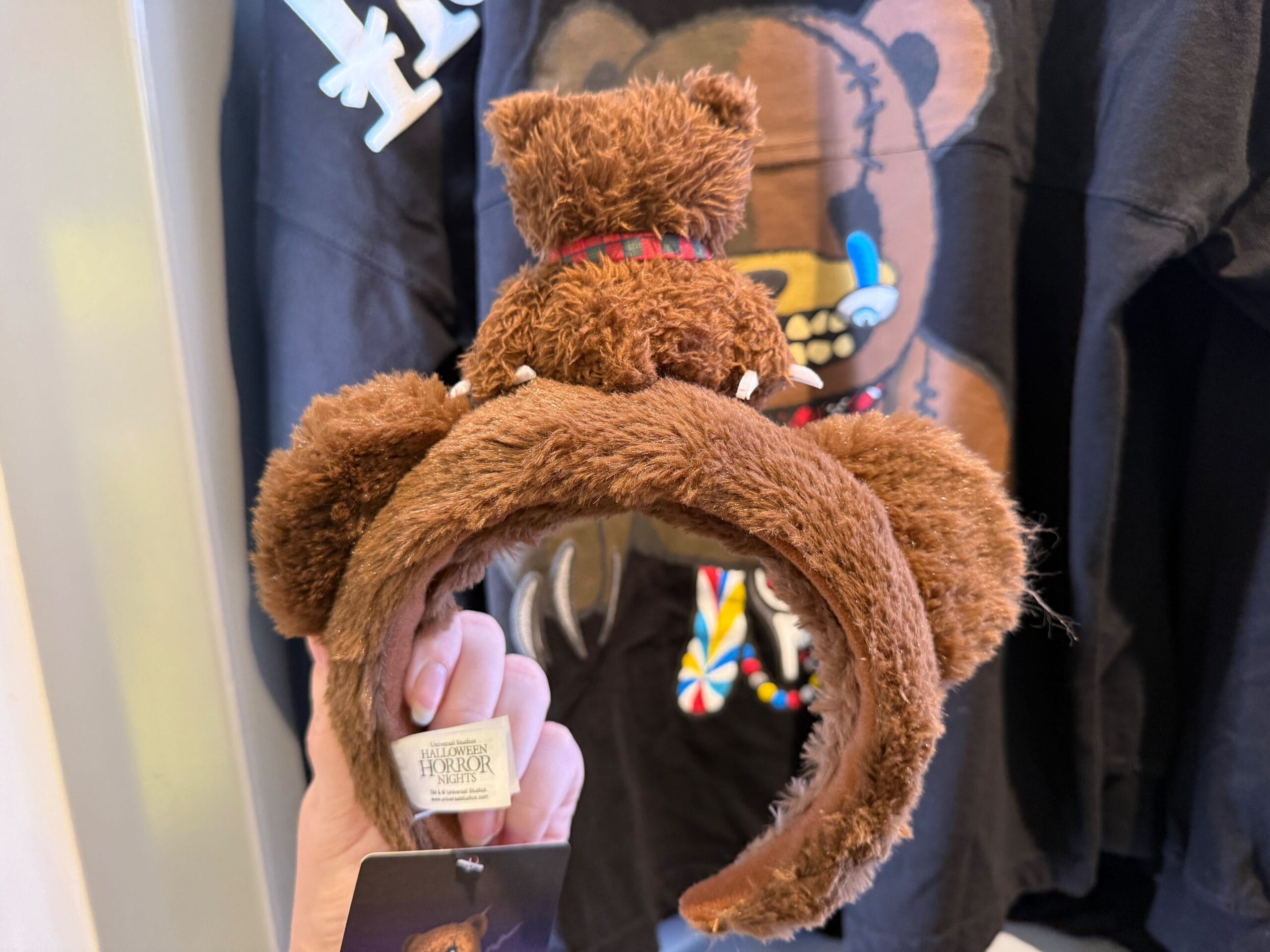 A hand holds a fluffy brown HamiKuma Merchandise bear ear headband before a black bear-themed shirt.