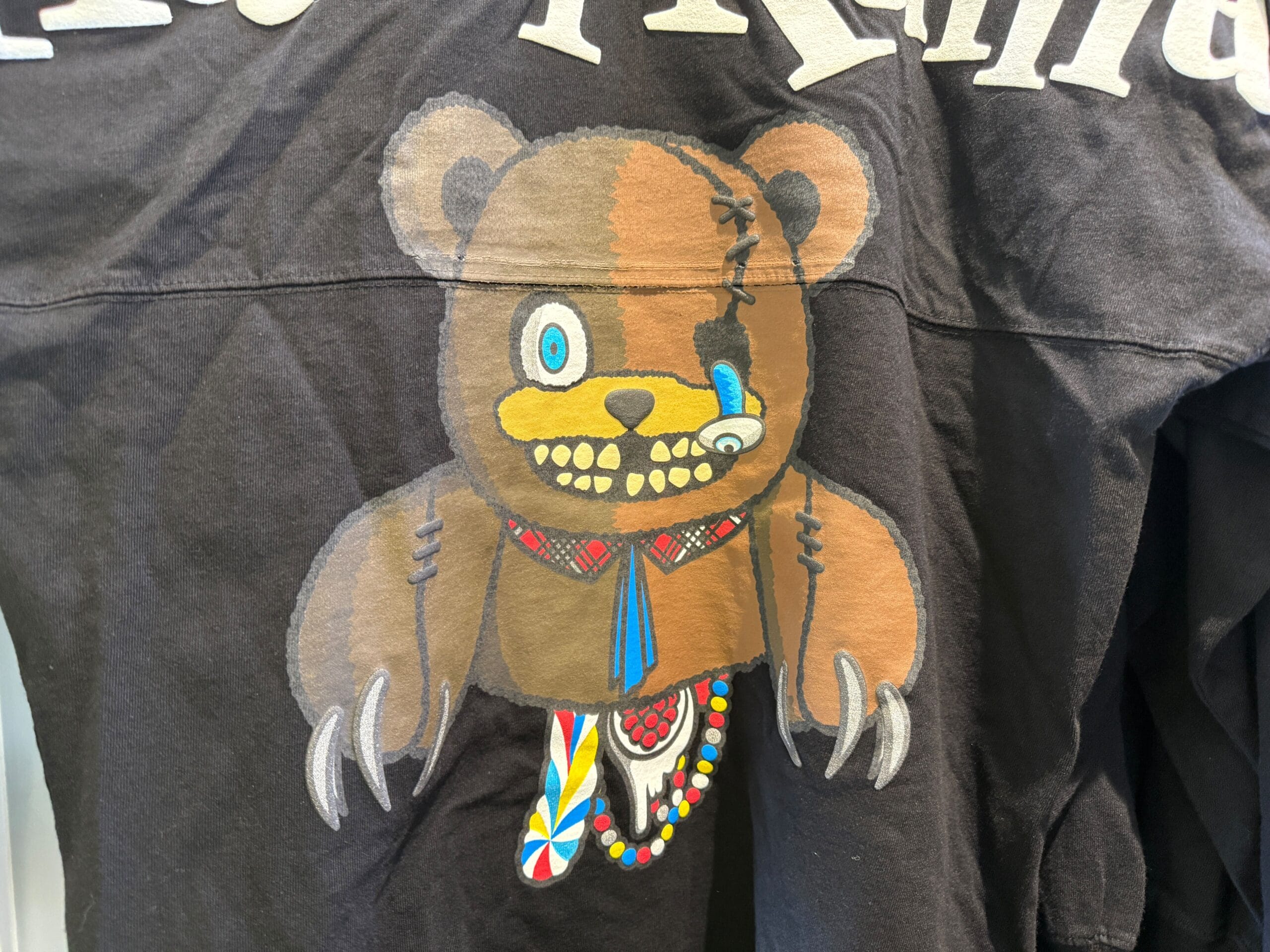 A cartoon bear with sharp claws, mismatched eyes, and colorful feathers on a HamiKuma Merchandise black shirt.