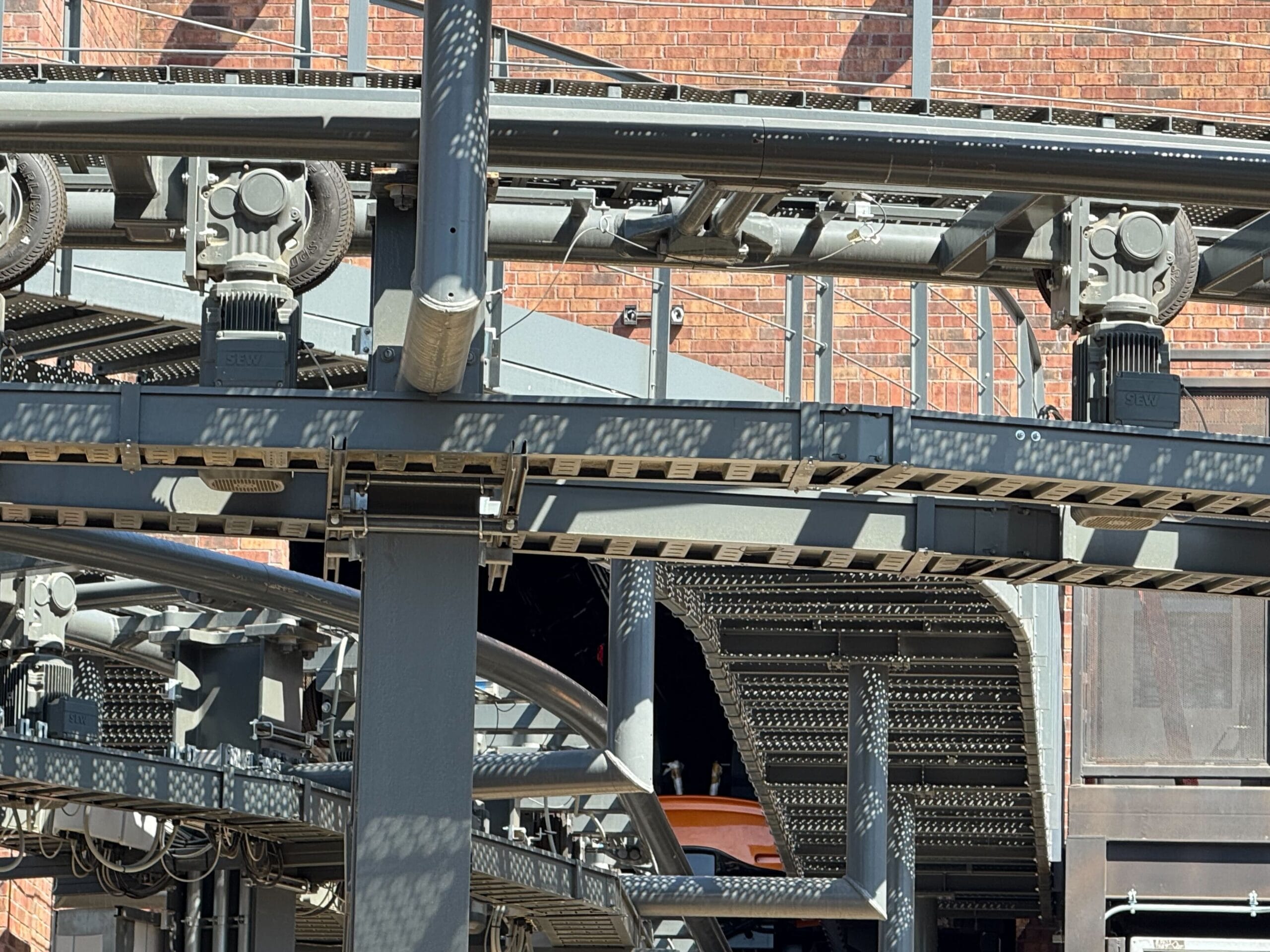 Close-up of a roller coaster track structure with metal supports and a brick building in the background.