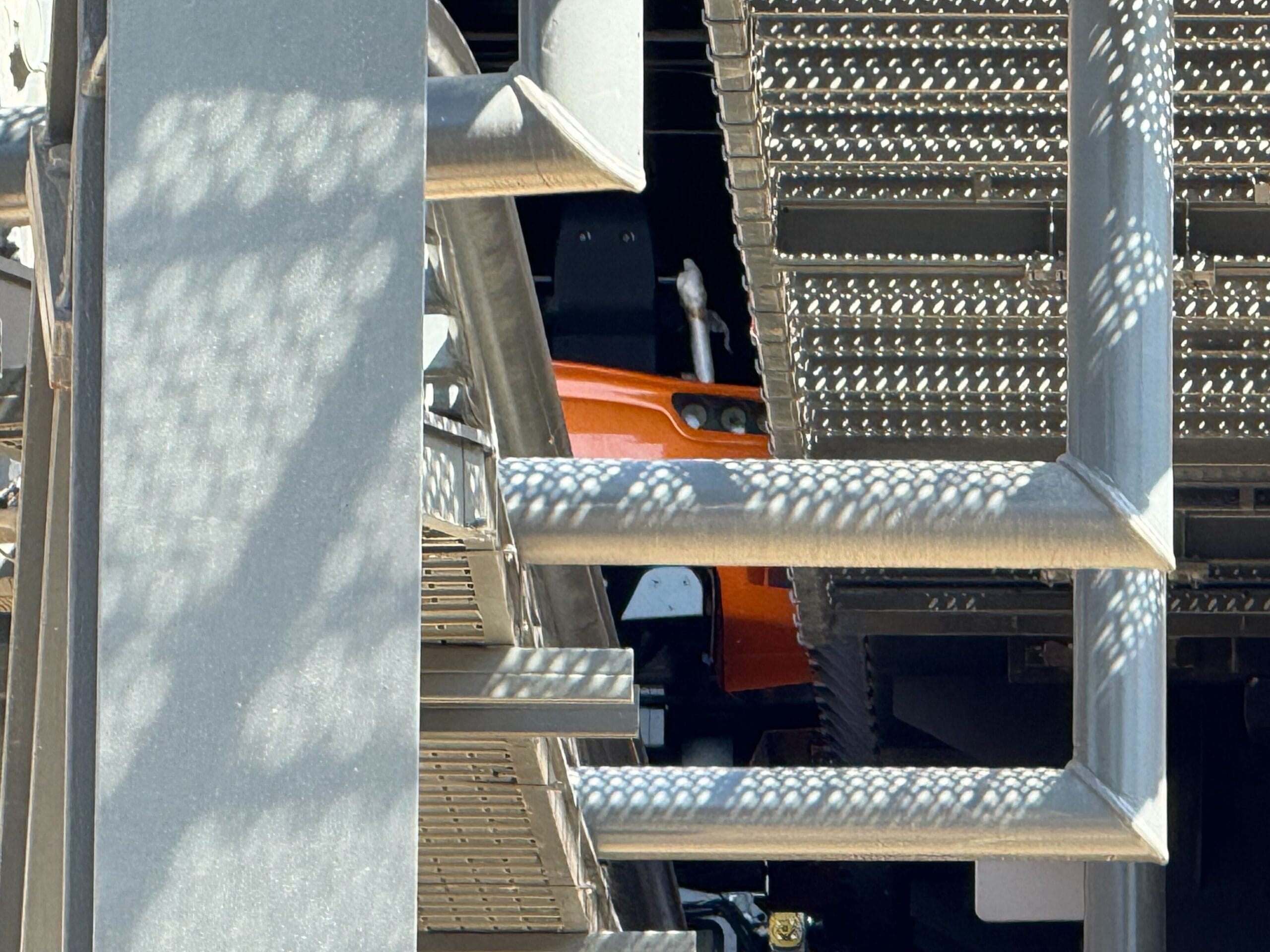 Close-up view of metal beams and grated platforms with a glimpse of orange machinery partially visible behind them.