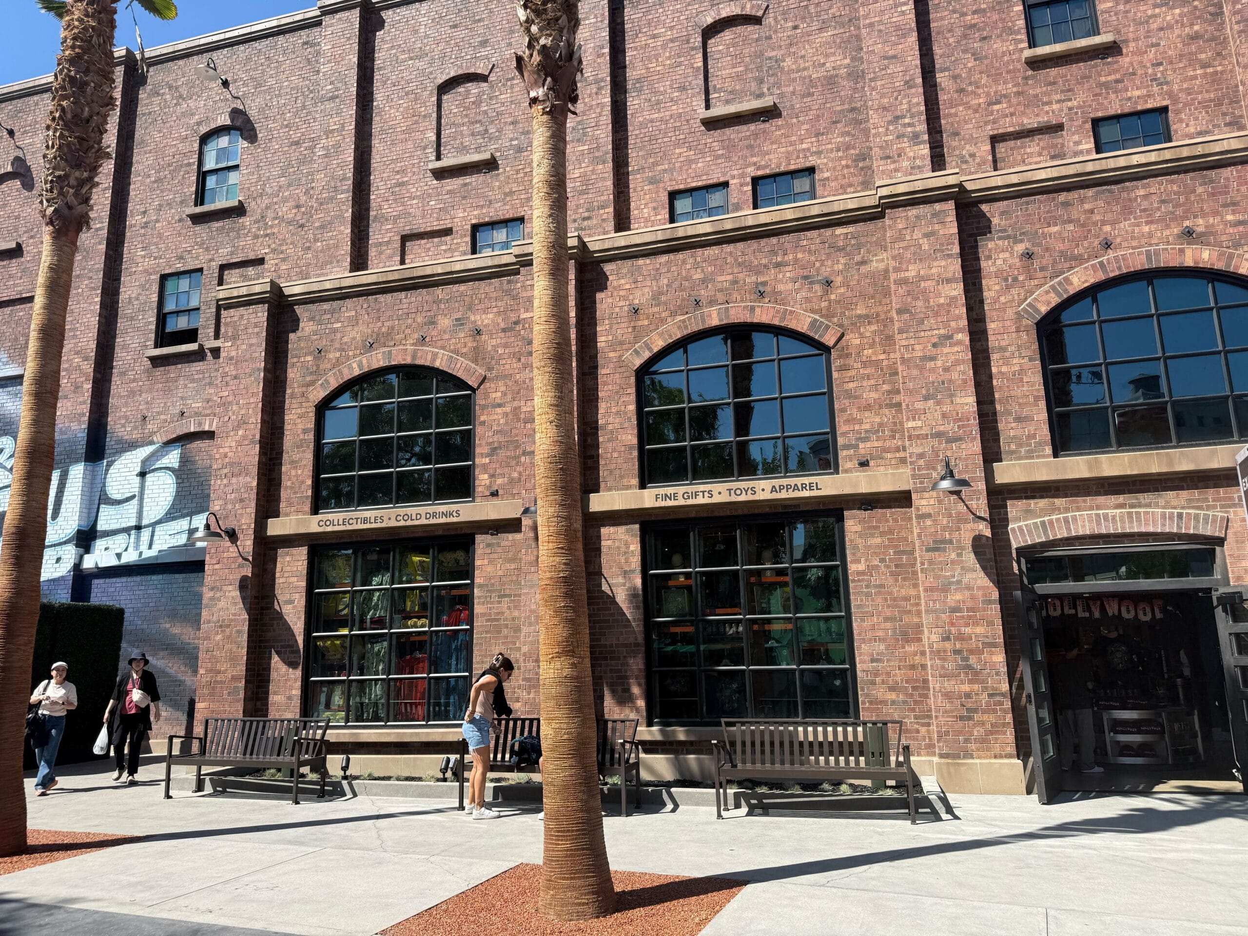 Brick building with large windows, palm trees, benches, and people walking—Hollywood Drift signs: "Fine Gifts" & "Cold Drinks.