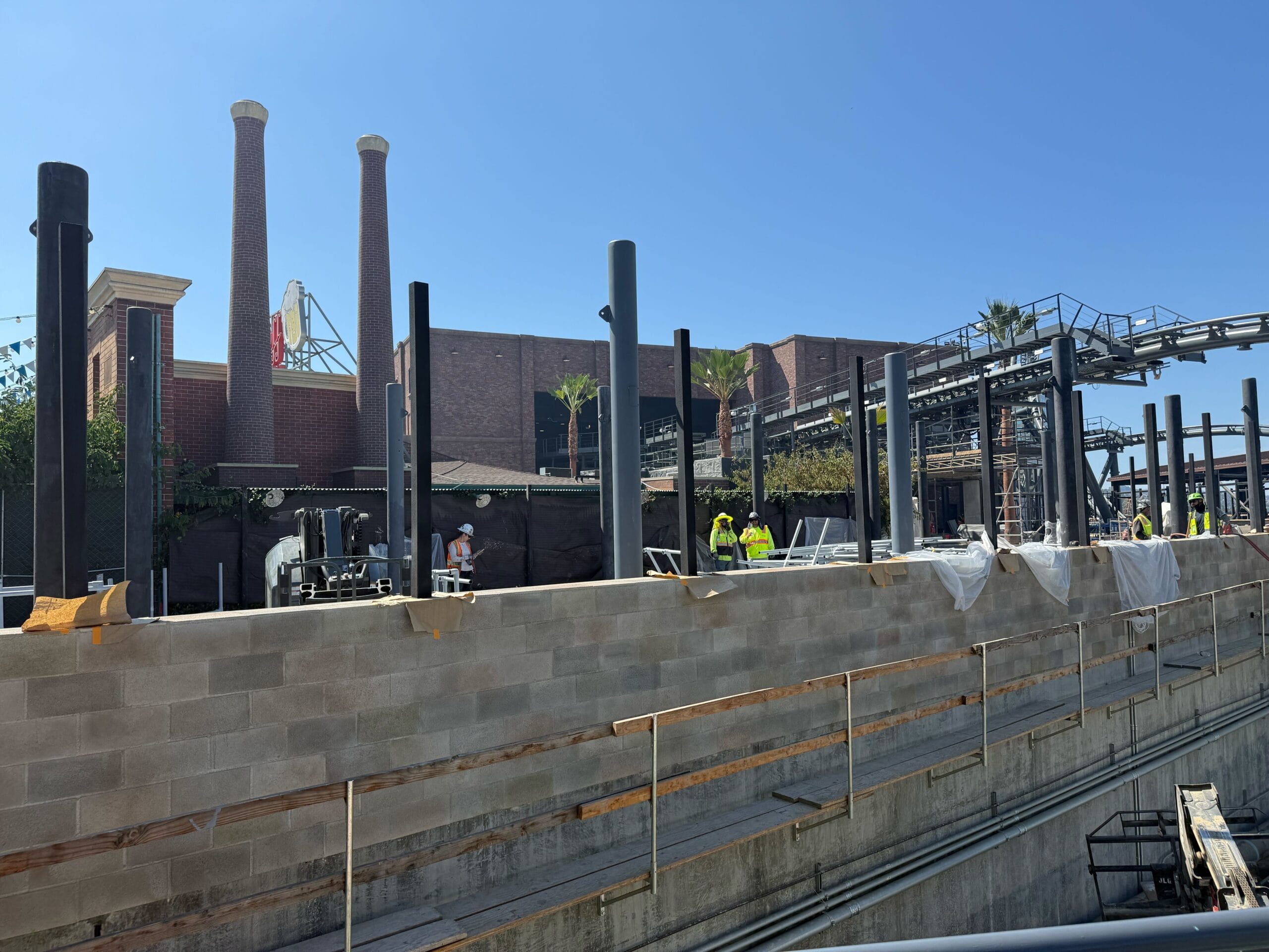 Construction workers build with steel beams and block walls, as Hollywood Drift echoes near a brick building with smokestacks.