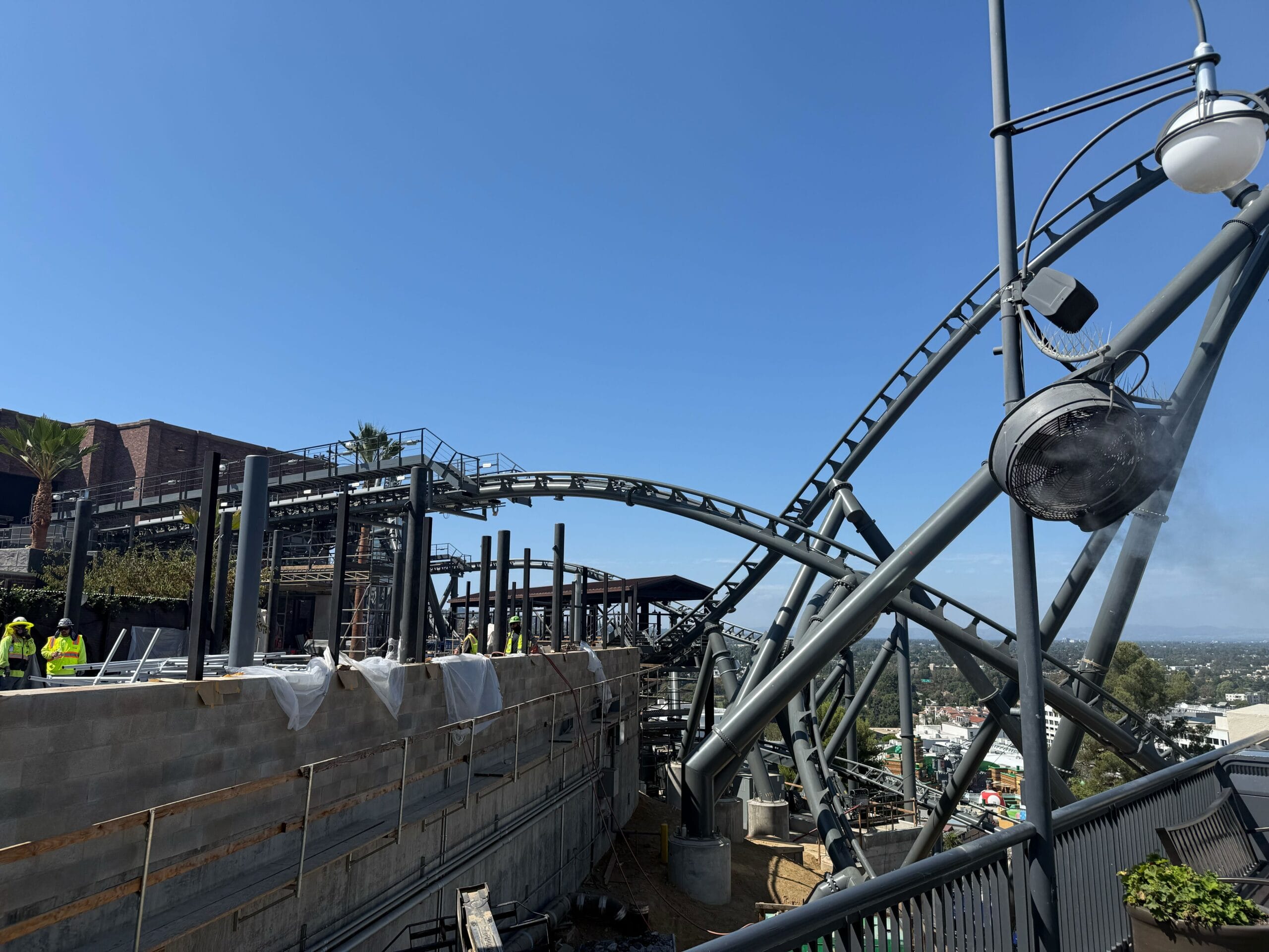 Construction workers build near the Hollywood Drift roller coaster under a clear blue sky, cityscape in the background.