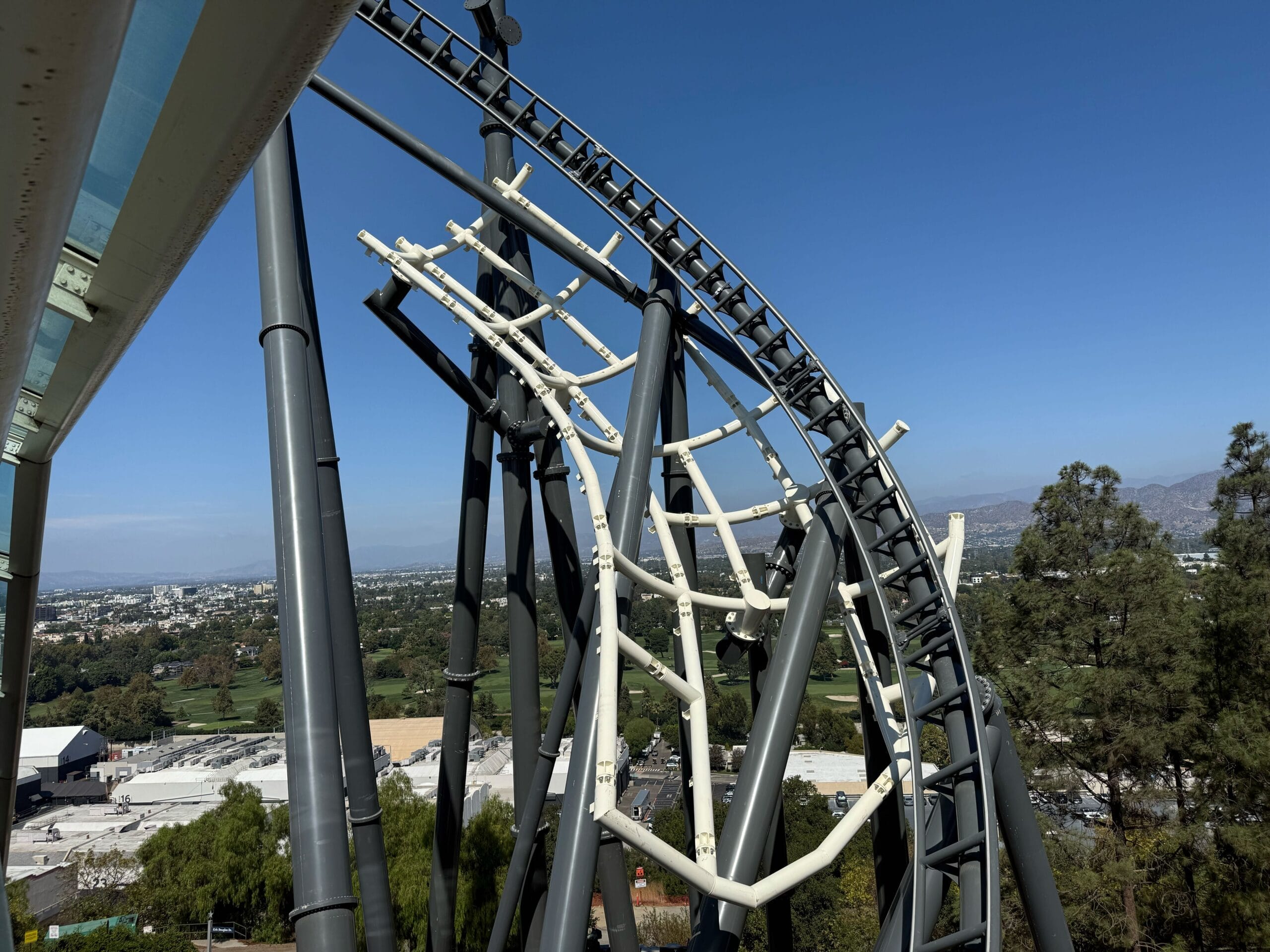 Close-up of the Hollywood Drift roller coaster track, with support beams silhouetted against a clear blue sky and city backdrop.