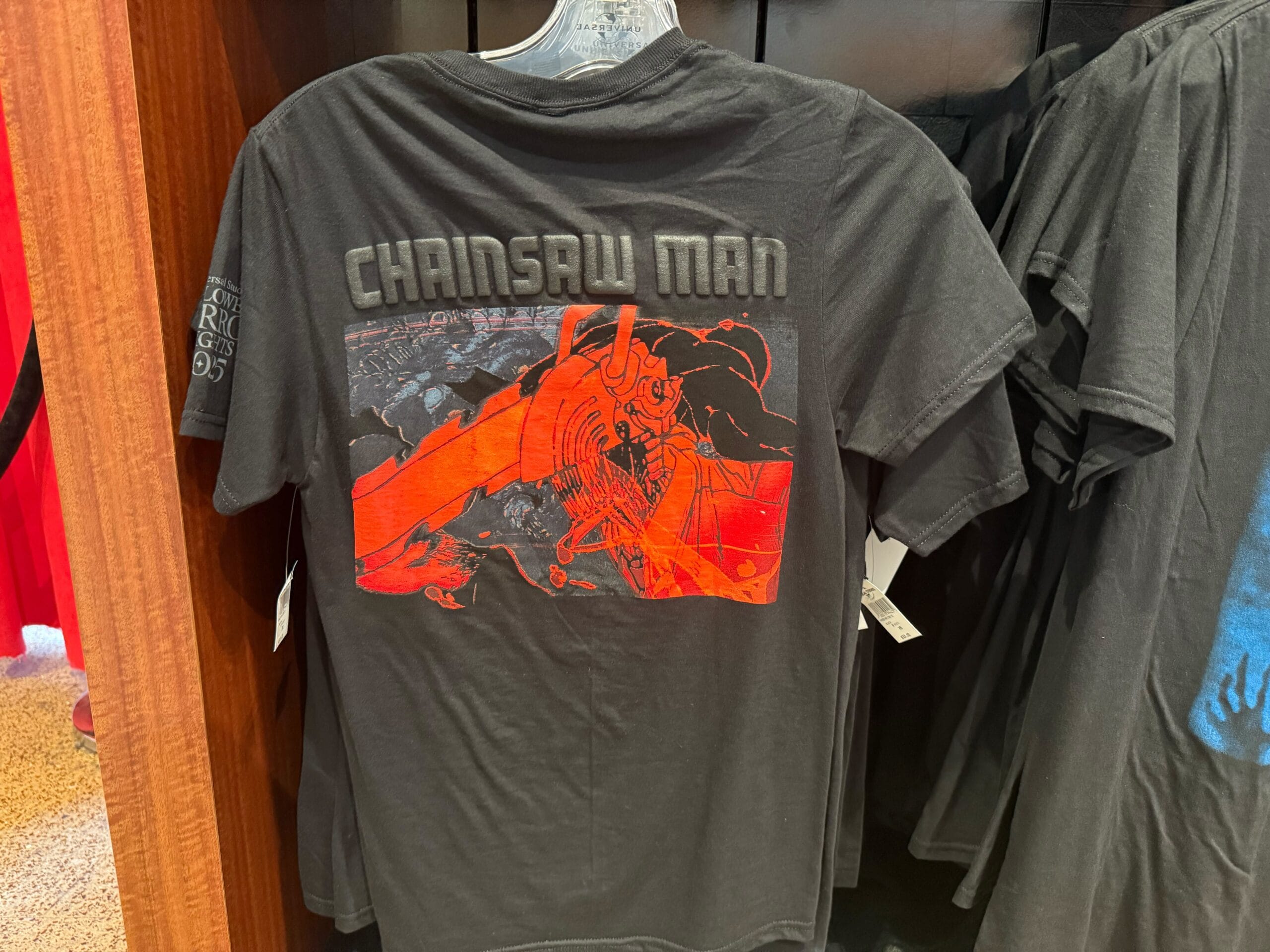 A black t-shirt featuring a red Chainsaw Man graphic, bold text, and exclusive HHN Merch 2025 style.