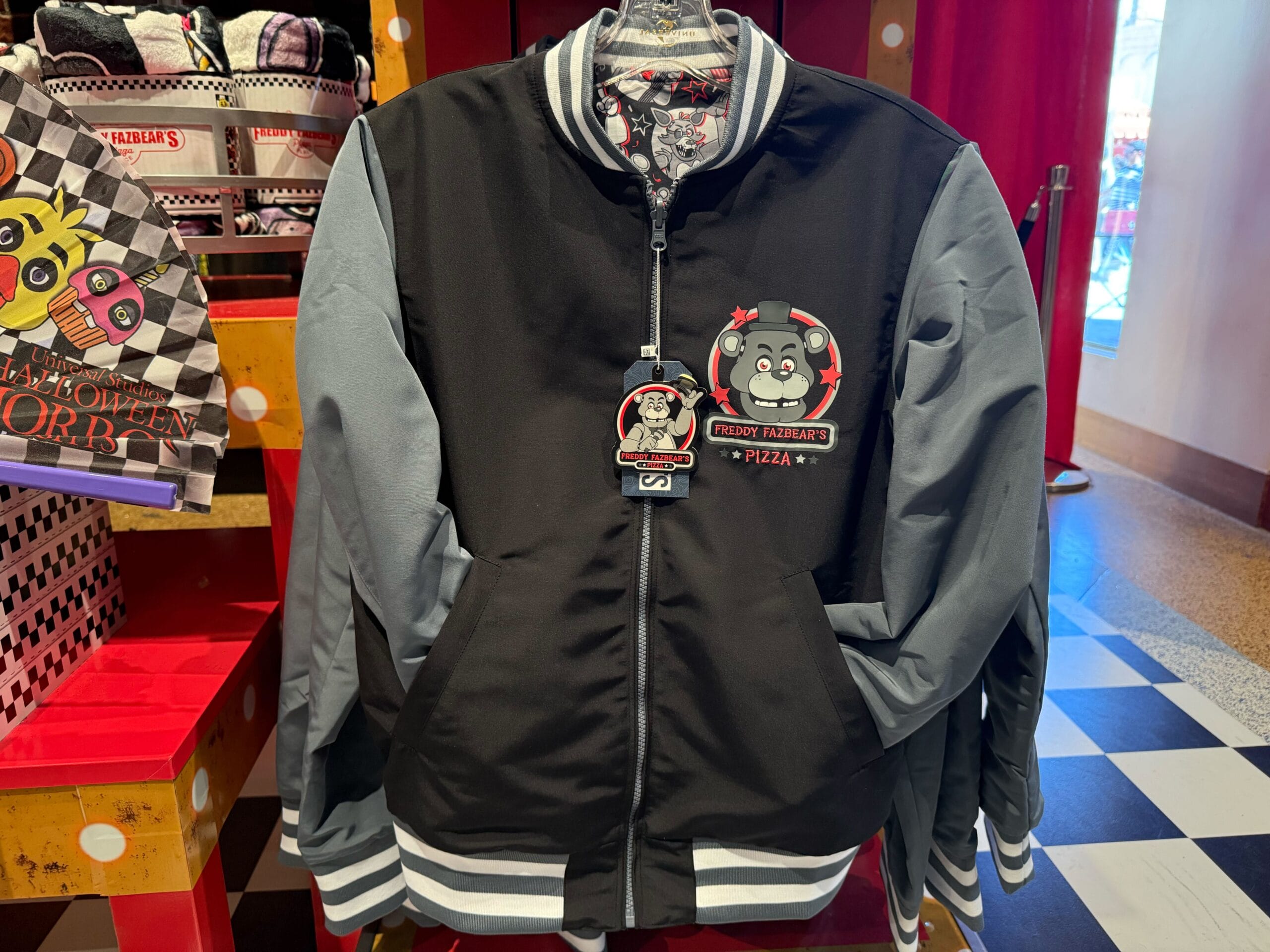 Black and gray jacket featuring a cartoon pizza mascot and “Freddy Fazbear's Pizza” logo, part of HHN Merch 2025.