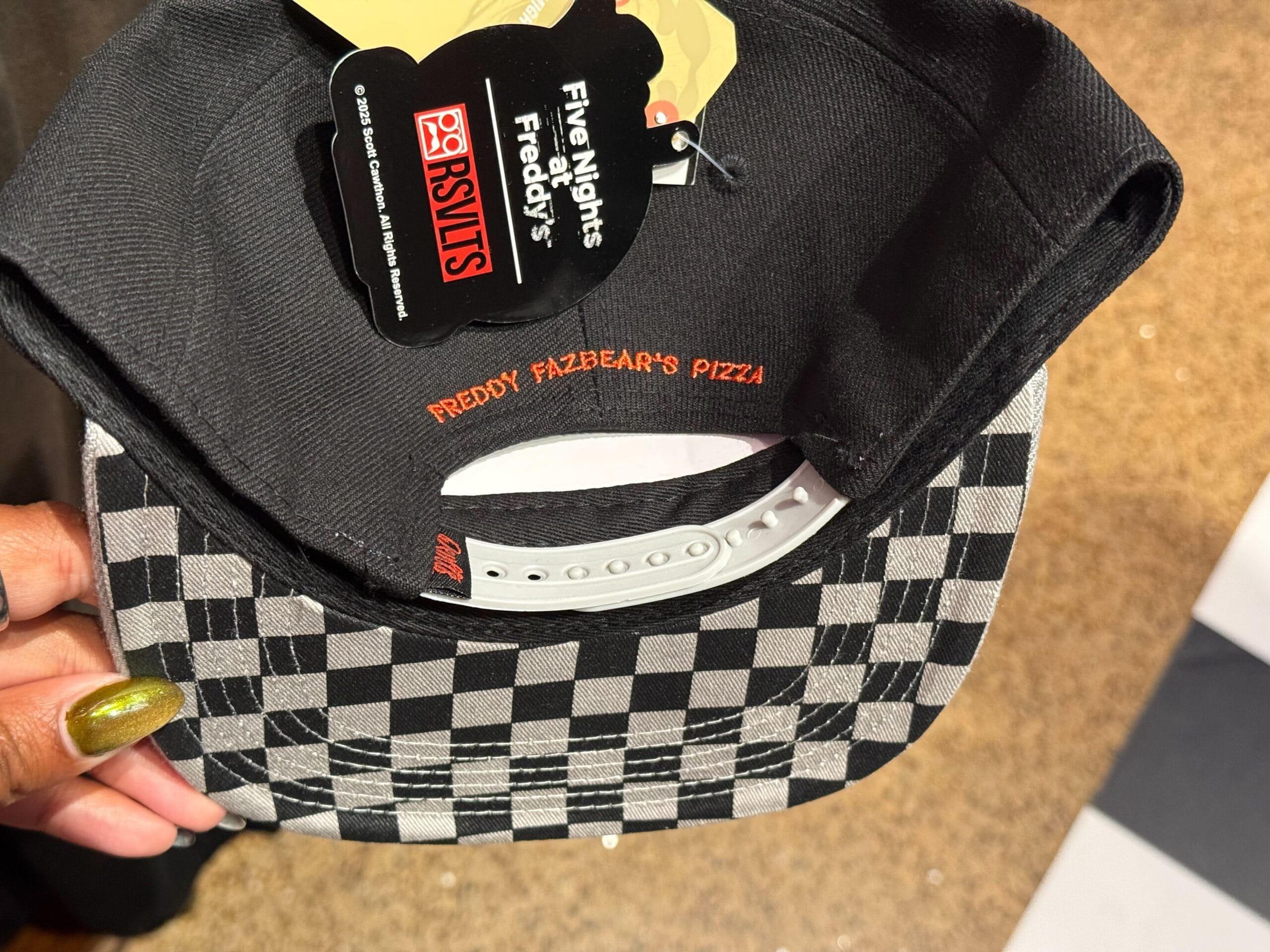 A hand holds a black cap with a checkerboard brim, part of the exclusive HHN Merch 2025 at Universal Studios Hollywood.
