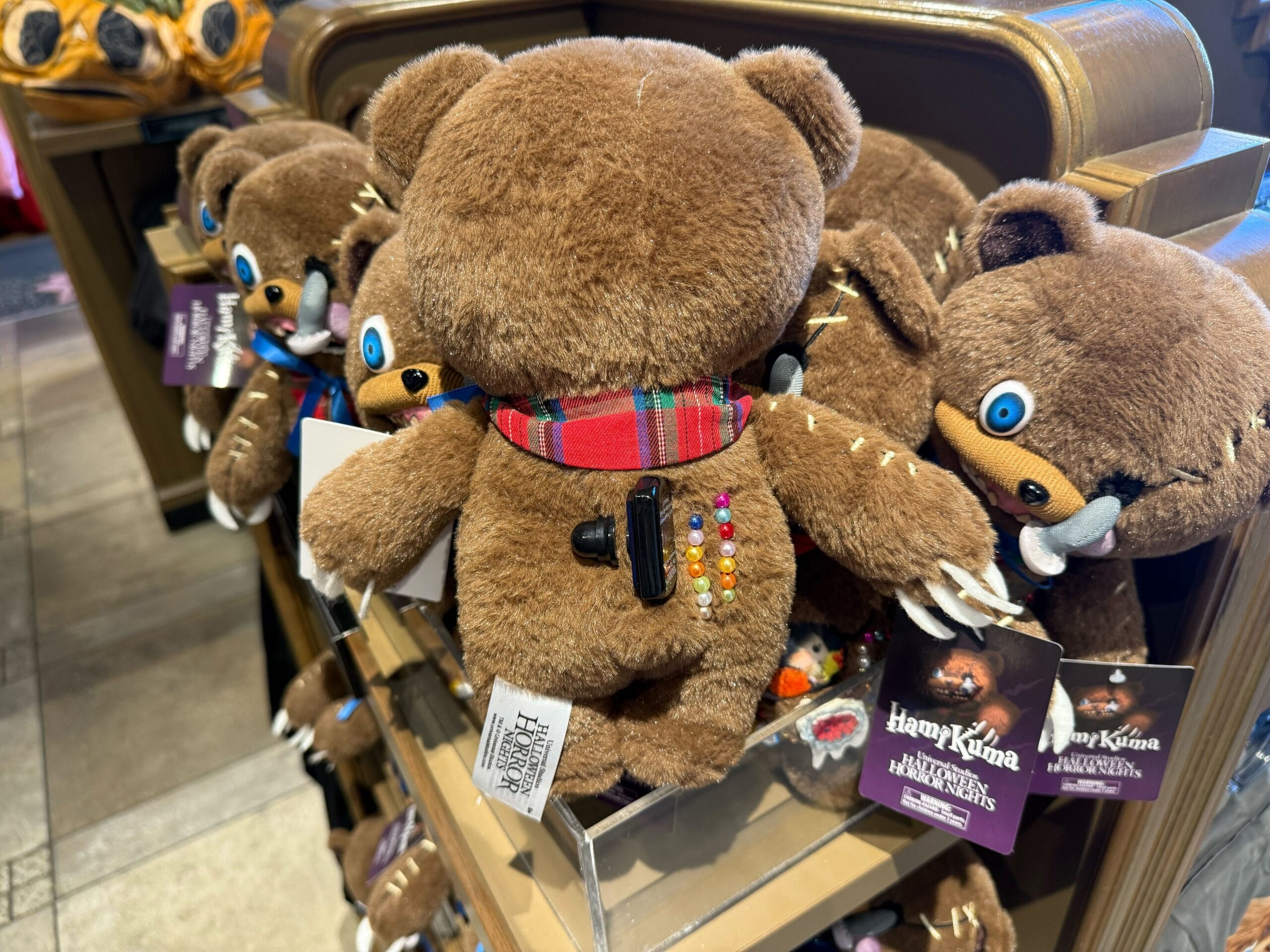 A plush bear with colorful pins and plaid scarf sits among Universal Studios Hollywood Halloween Horror Nights merchandise.