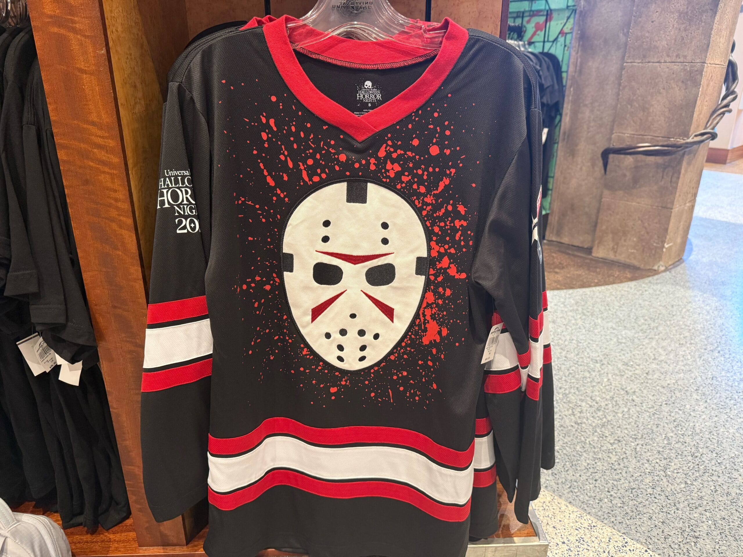 Black hockey jersey with a white and red goalie mask, splatter accents, and bold stripes—perfect HHN Merch 2025 style.