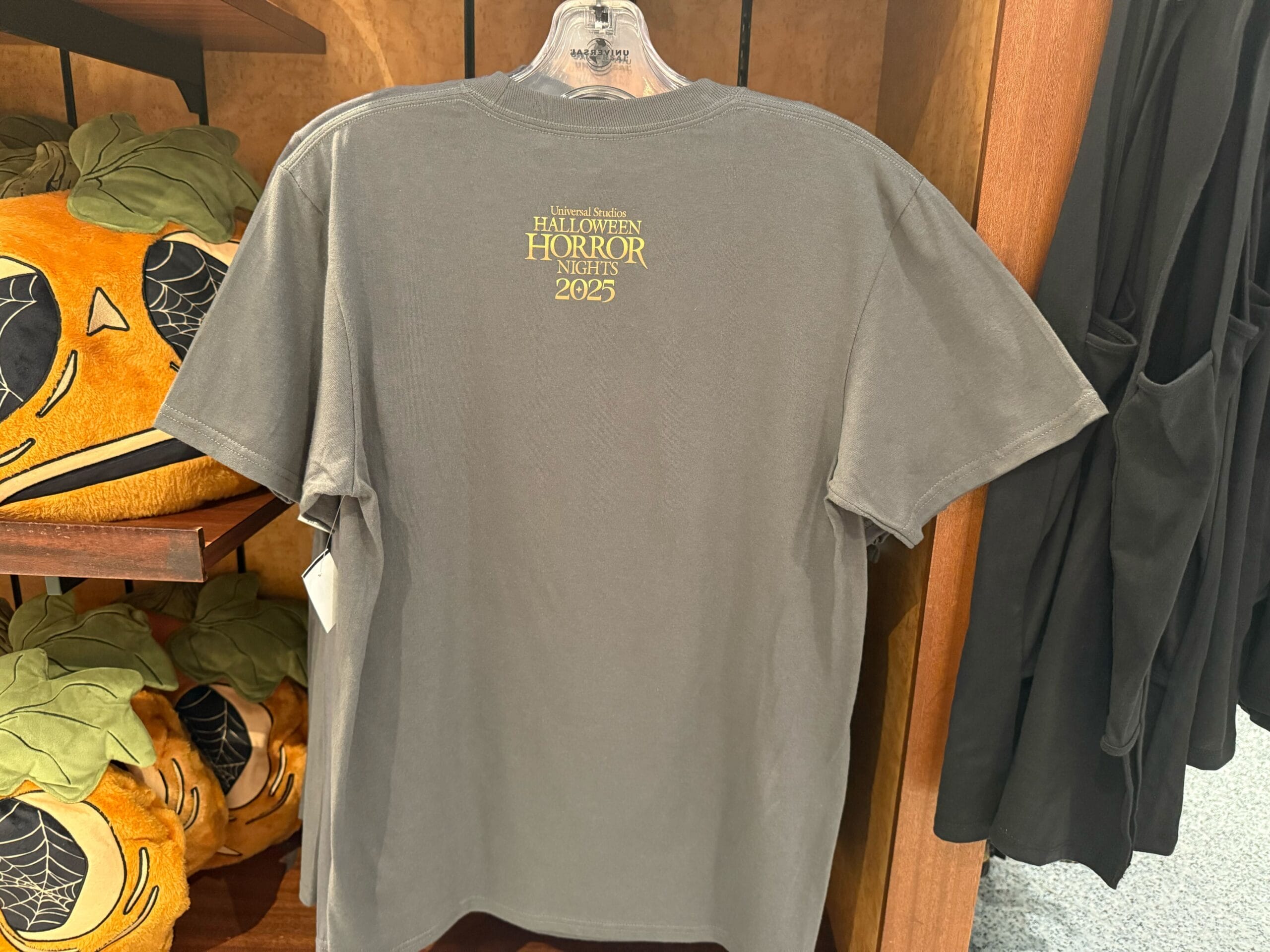 Gray T-shirt on a hanger with "HHN Merch 2025" text, next to pumpkin plushies at Universal Studios Hollywood.