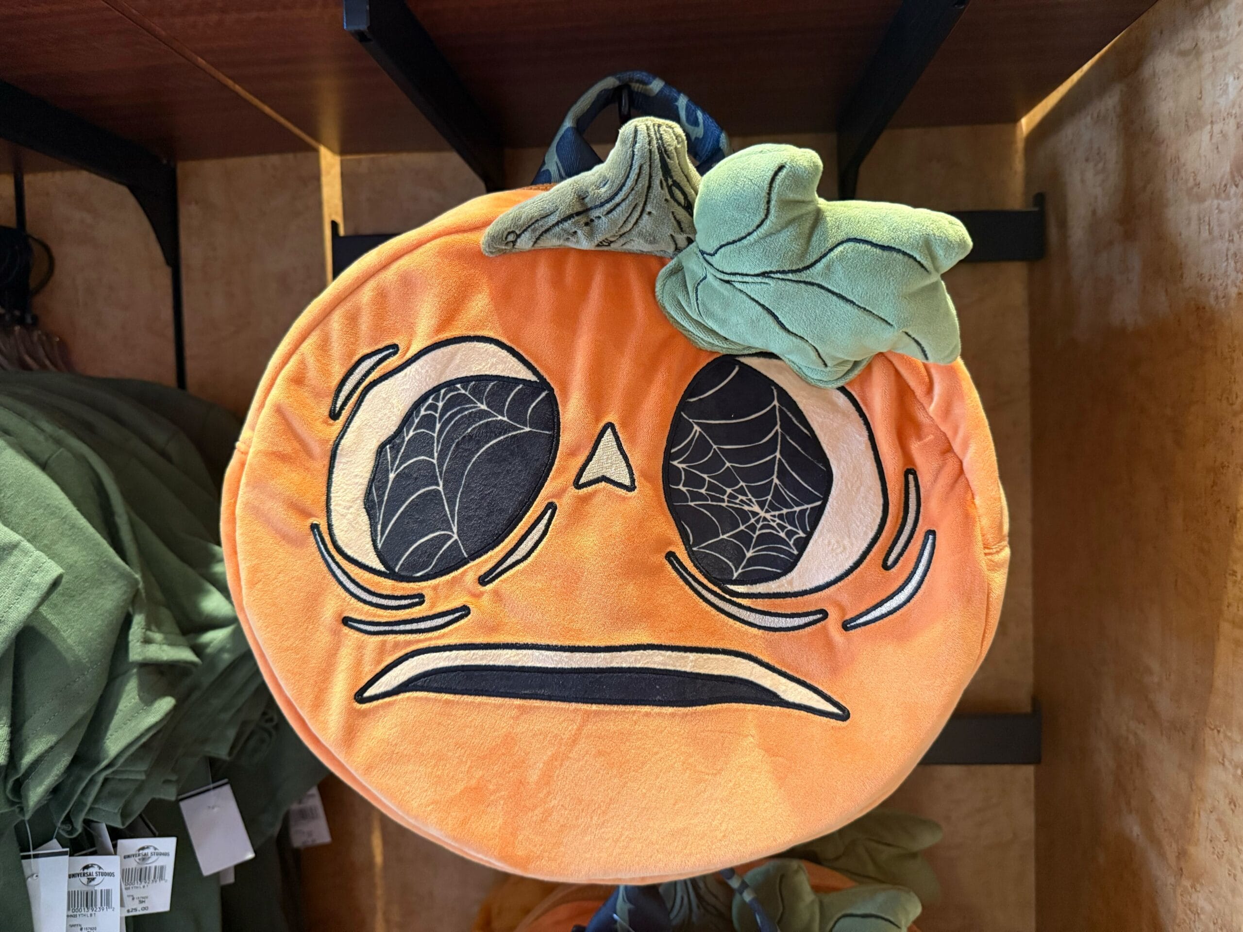 A plush pumpkin with spiderweb eyes and a worried expression hangs among Halloween Horror Nights Merchandise.